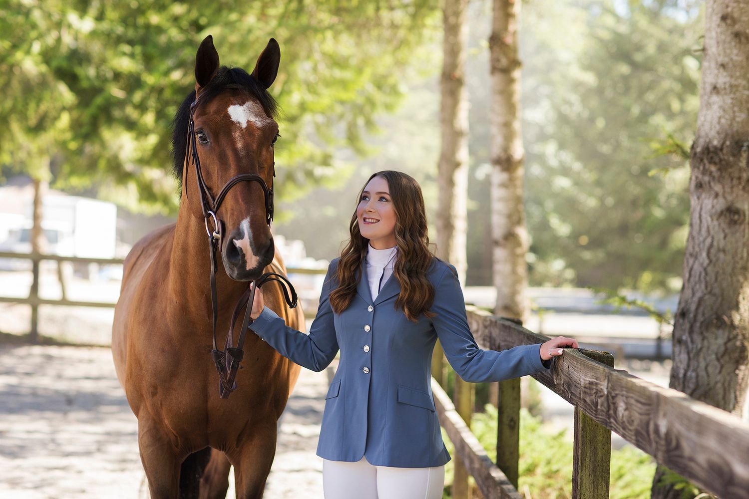Young woman in blue equestrian jacket smiling beside a chestnut horse at a sunny stable fence.
