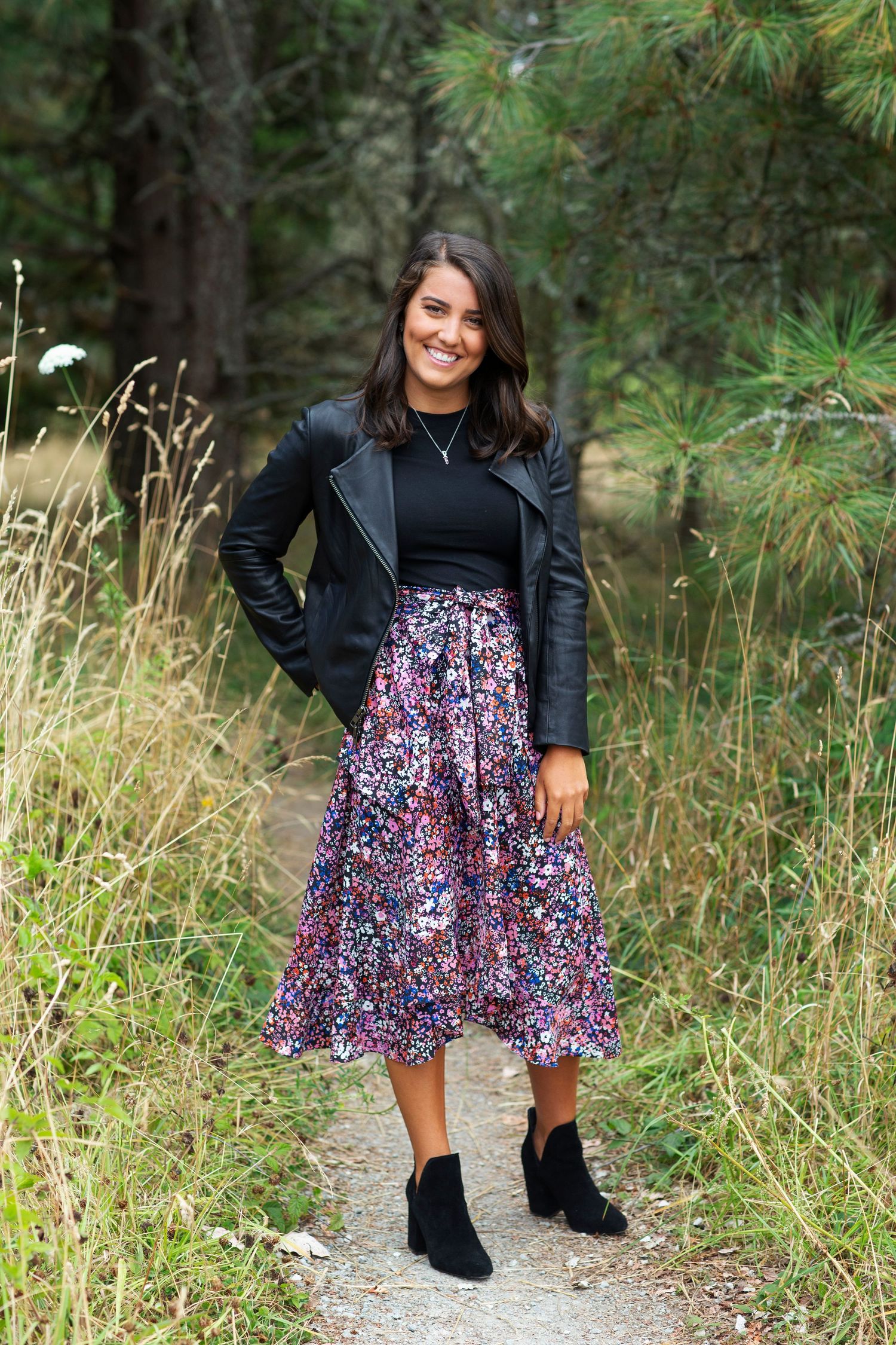 Stylish woman in leather jacket and floral skirt on forest trail.