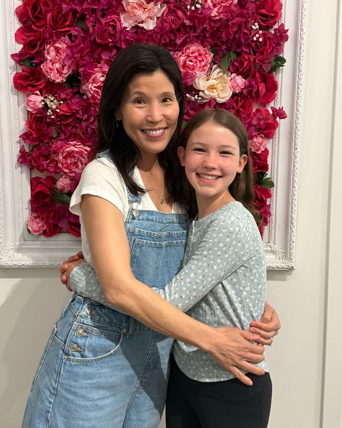 Woman in denim overalls hugs smiling girl in front of pink floral wall backdrop.