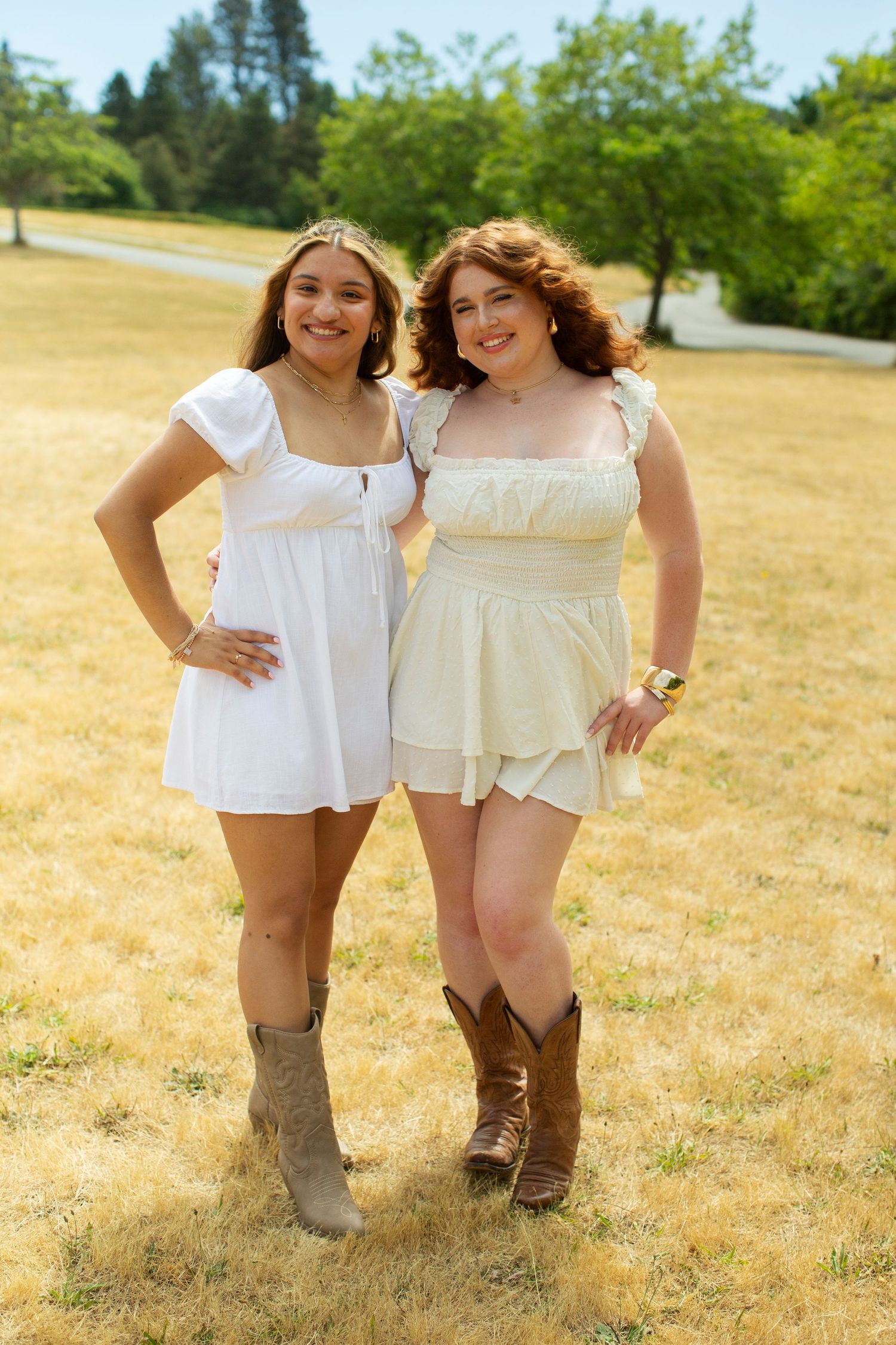 Two young women in white dresses smiling together in sunny park.