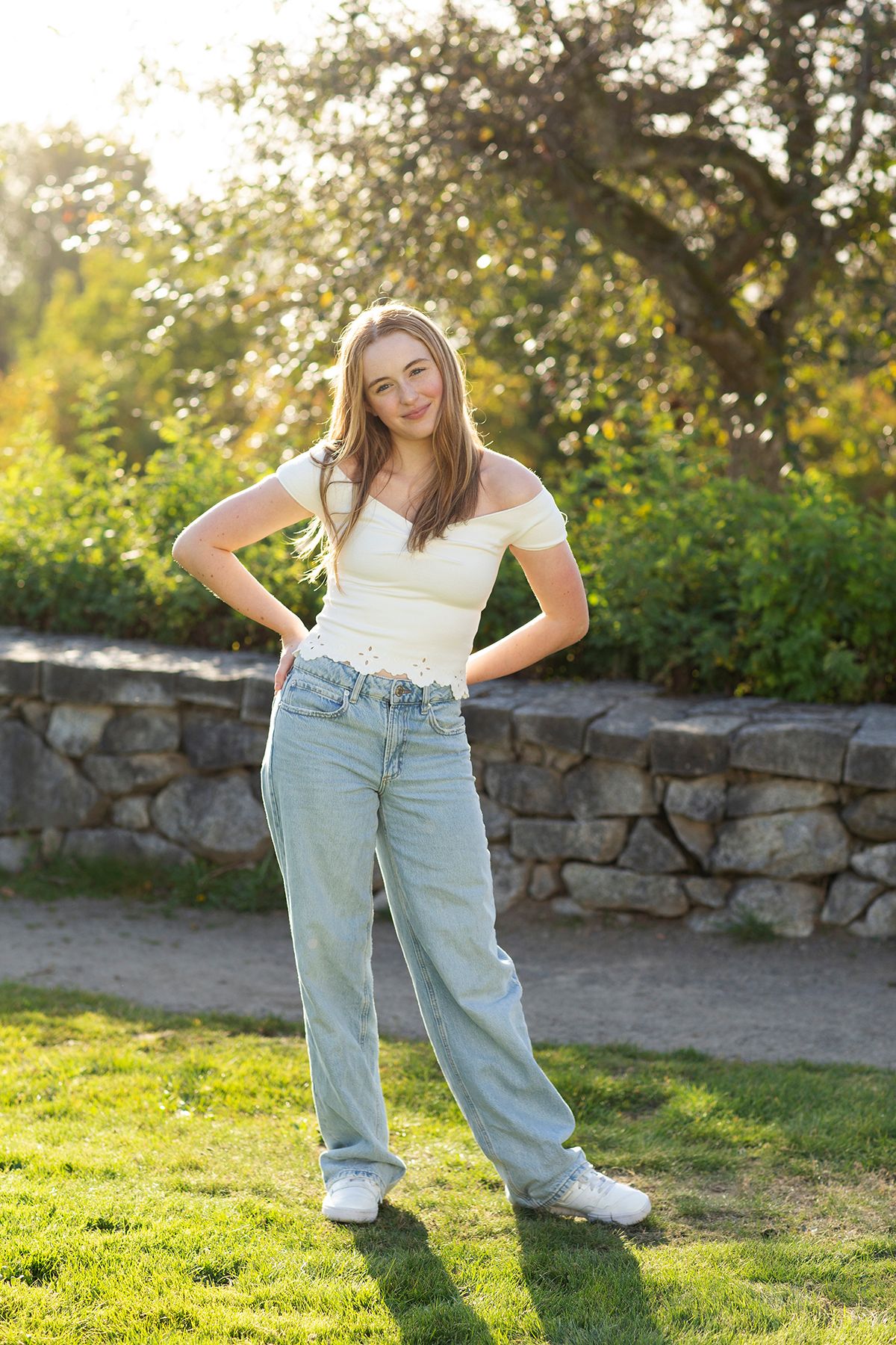 Young woman in white off-shoulder top and light blue jeans posing outdoors in a sunny garden with stone wall.