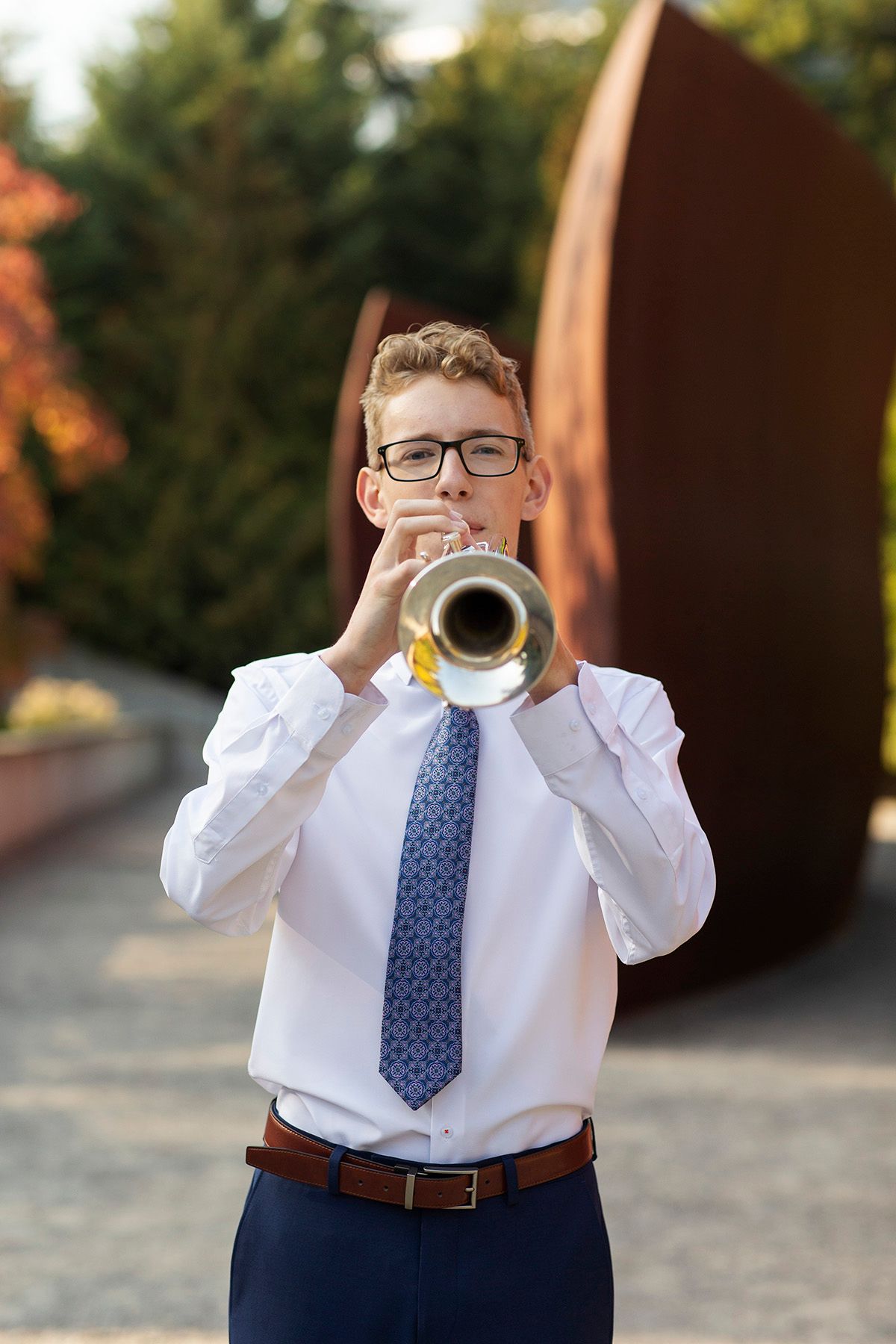 A young boy in glasses, white shirt, and blue tie plays trumpet outdoors near autumn trees and a sculpture.