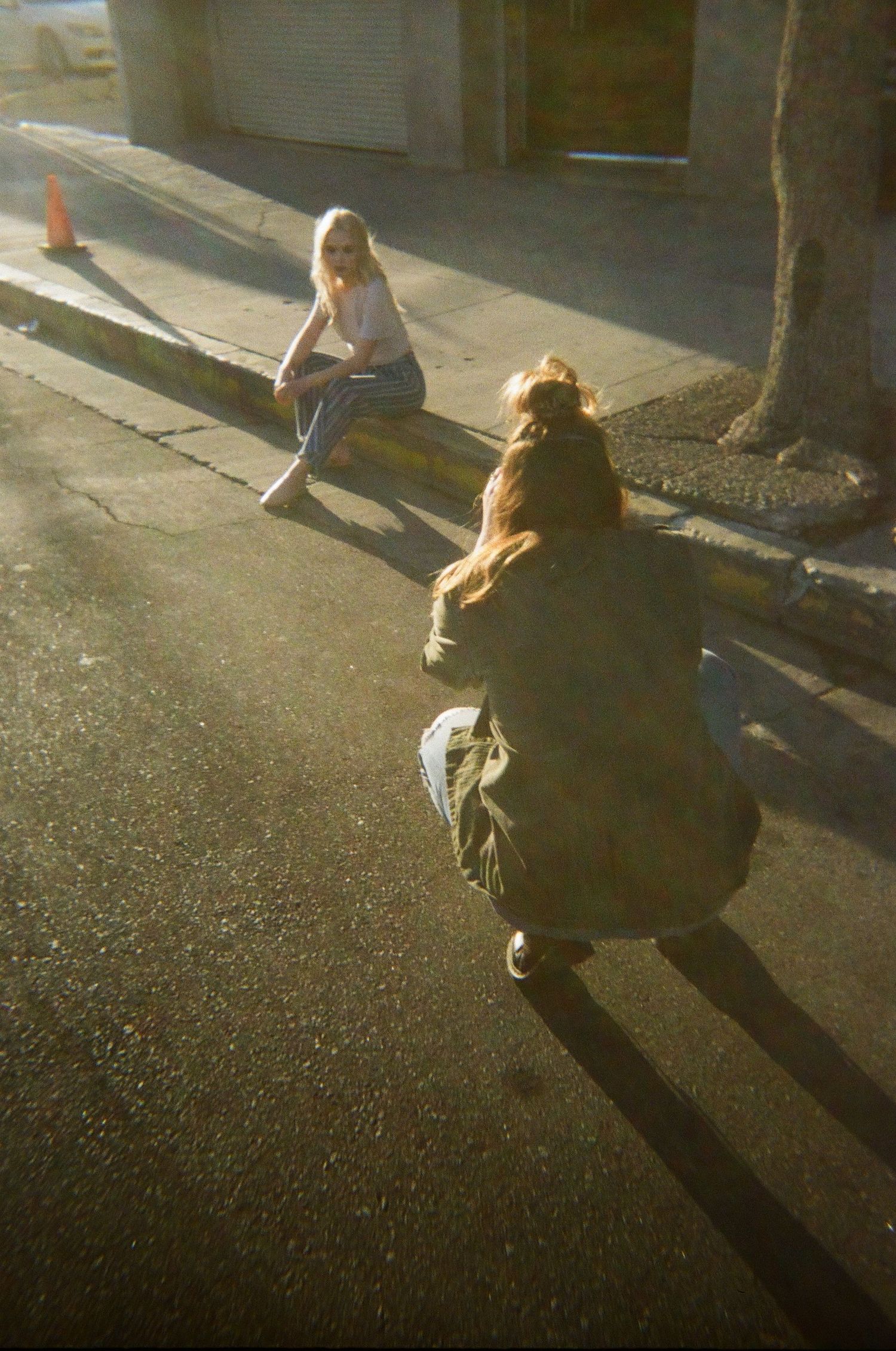 Two women sitting on curb in warm sunlight on urban street.