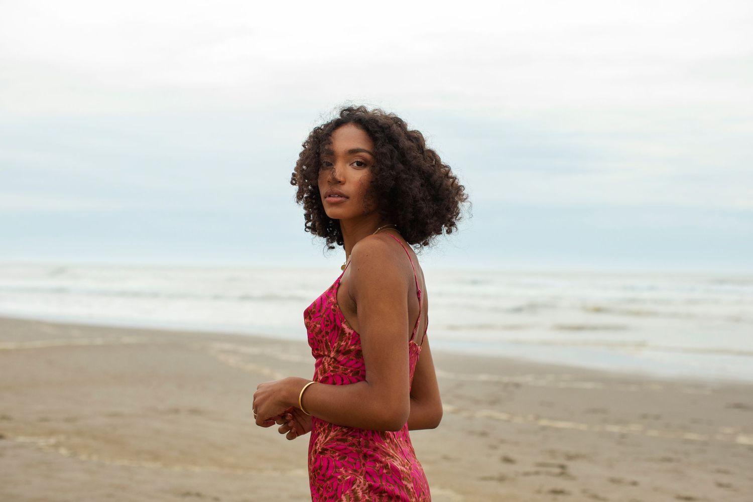 Young woman in a pink floral dress standing on a beach with ocean waves in the background.
