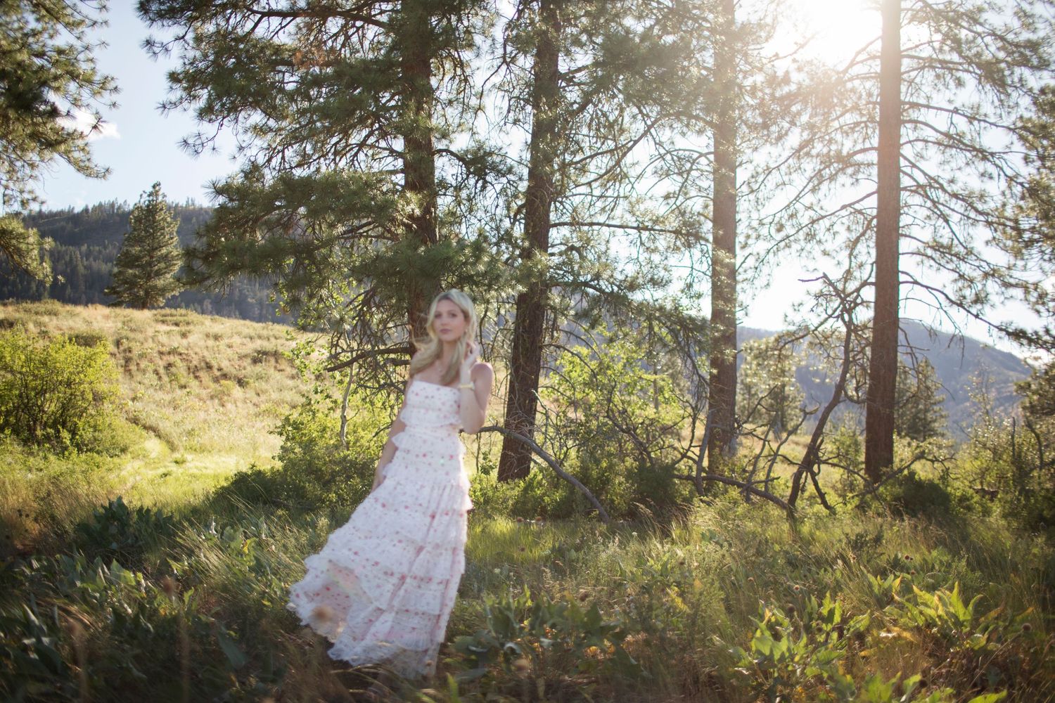 Woman in white floral dress standing among pine trees with golden sunlight streaming through the forest.