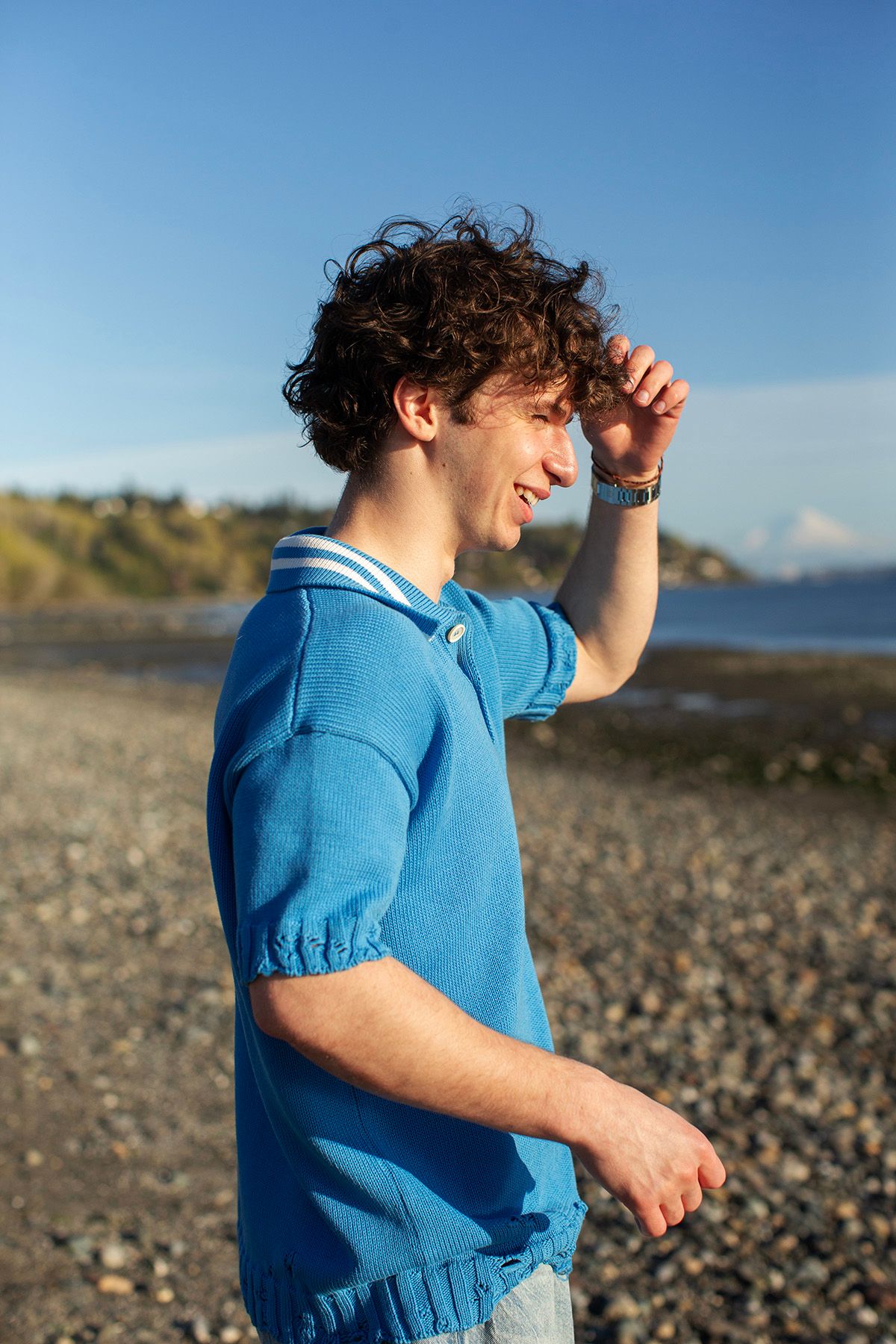 Young man with curly hair in blue polo shirt smiling on rocky beach with blue sky background.