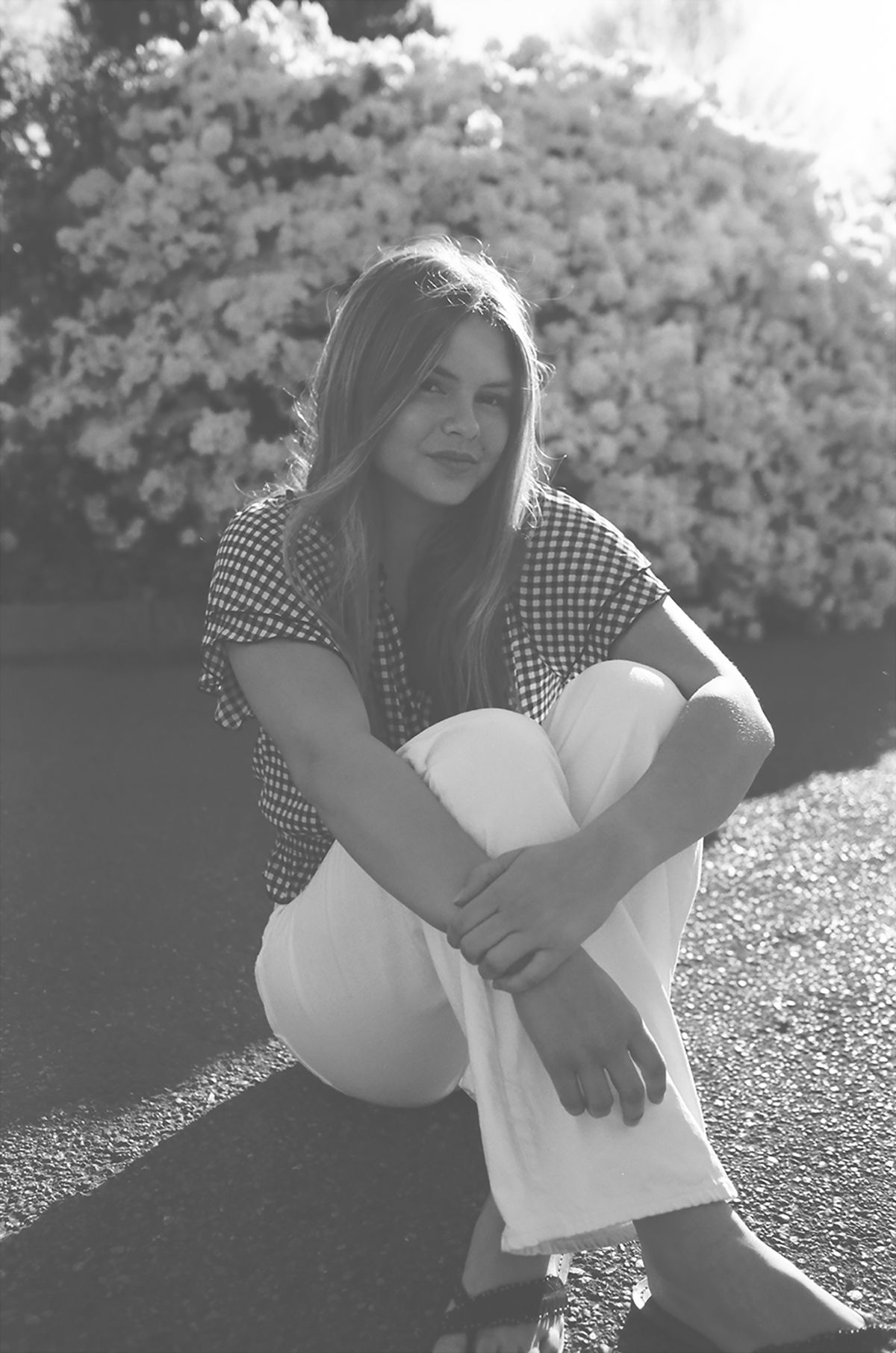 Black and white photo of a young woman in checkered top and white pants sitting outdoors near blooming bushes.