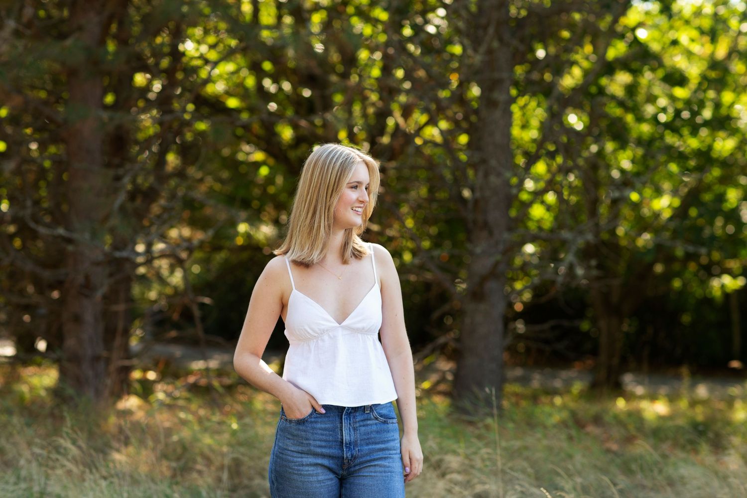 Blonde young woman in white top and jeans standing in wooded area.