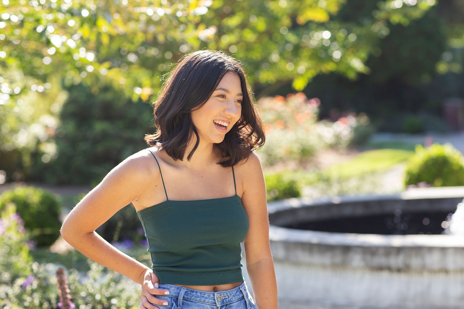 Young woman in green tank top and jeans smiling outdoors near a fountain in a sunny garden.