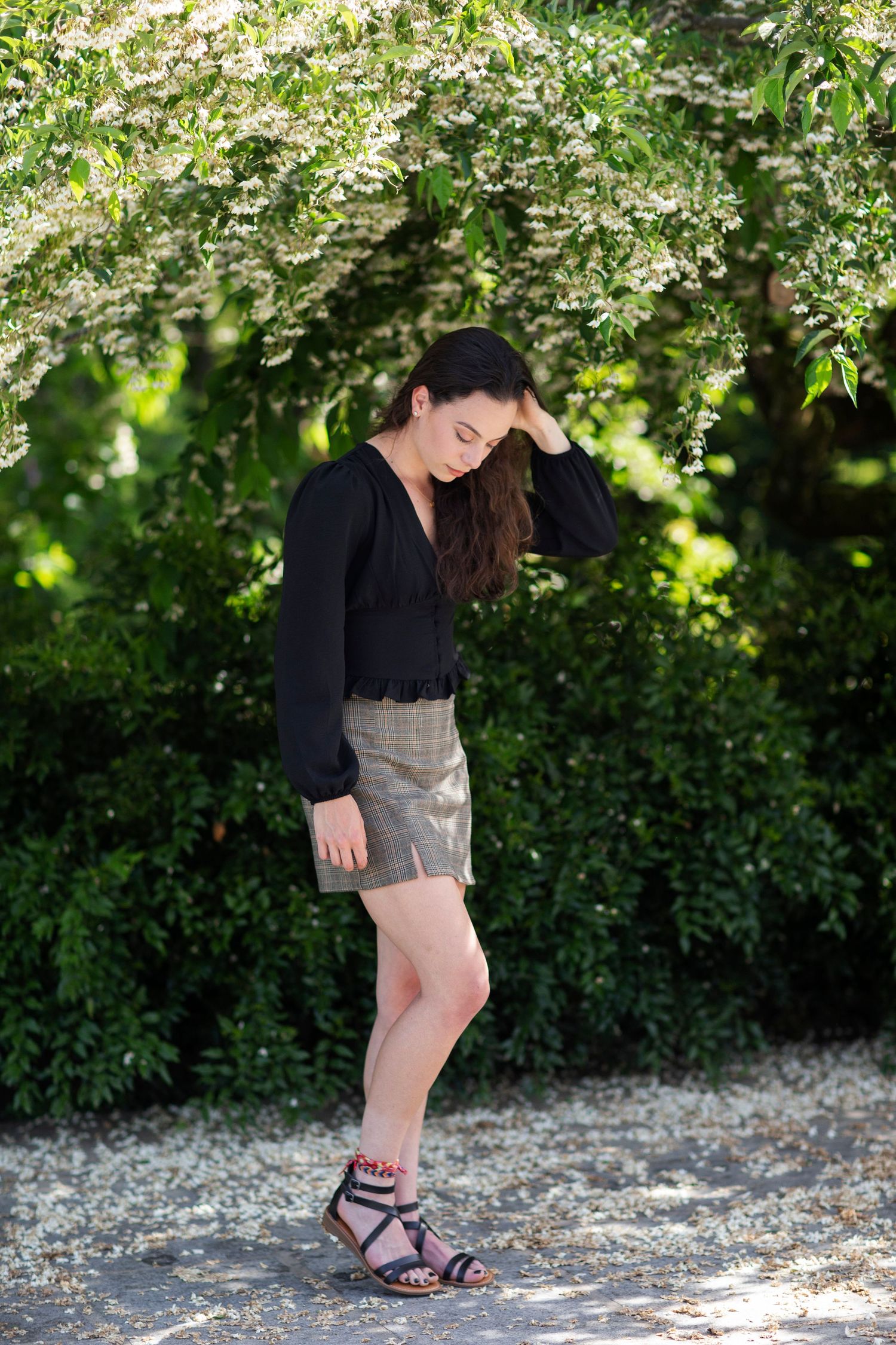 Young woman in black top and plaid shorts poses gracefully among blooming white flowers and lush green foliage outdoors.