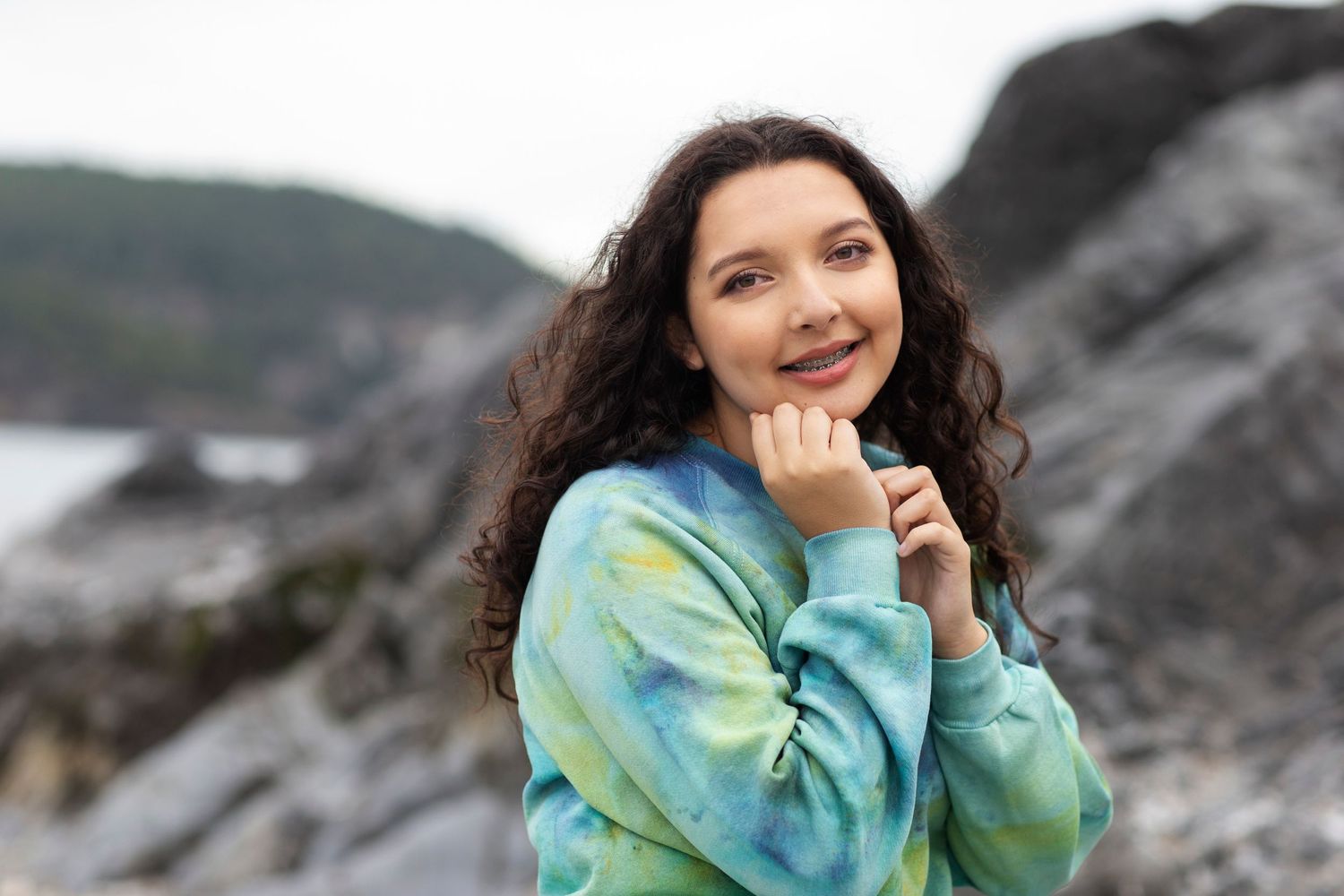 Young woman in tie-dye blue sweatshirt smiling with rocky coastal cliffs in the background.