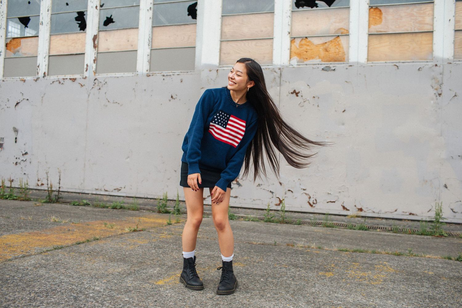 Young woman in American flag sweater laughing outdoors, long hair flowing, posing near weathered industrial building.