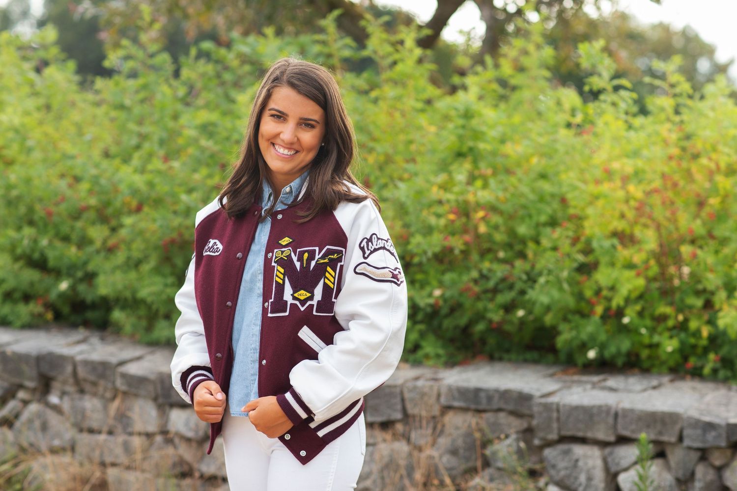 Young woman smiling outdoors wearing a maroon and white varsity letterman jacket with M logo.