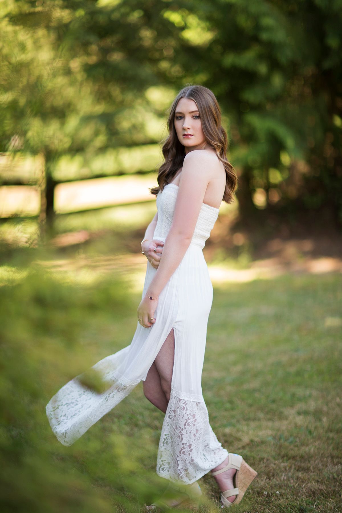 Young woman in white lace gown posing outdoors in a green park setting, looking over her shoulder.