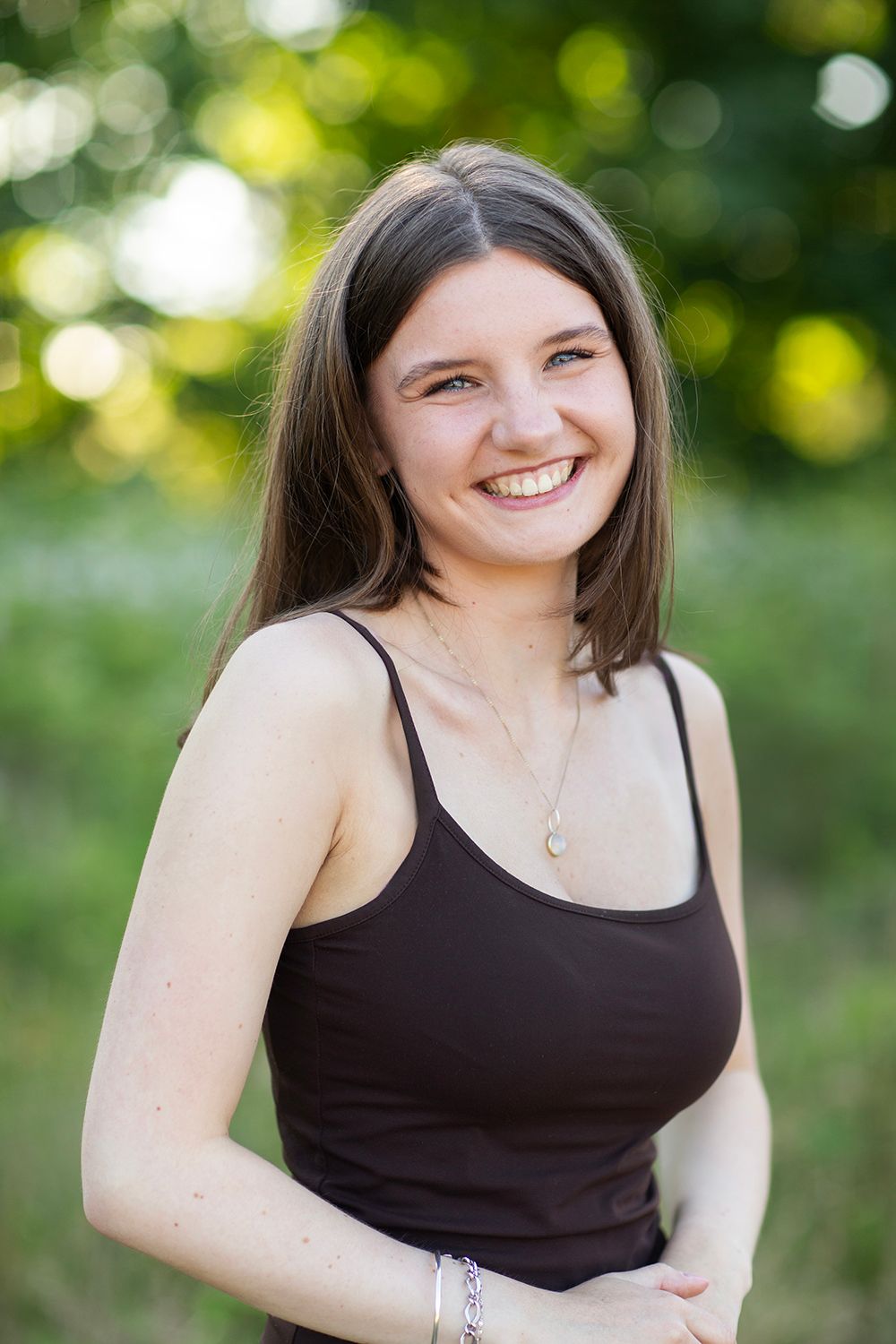 Smiling young woman with brown hair wearing a dark tank top and necklace, posing outdoors with green foliage background.
