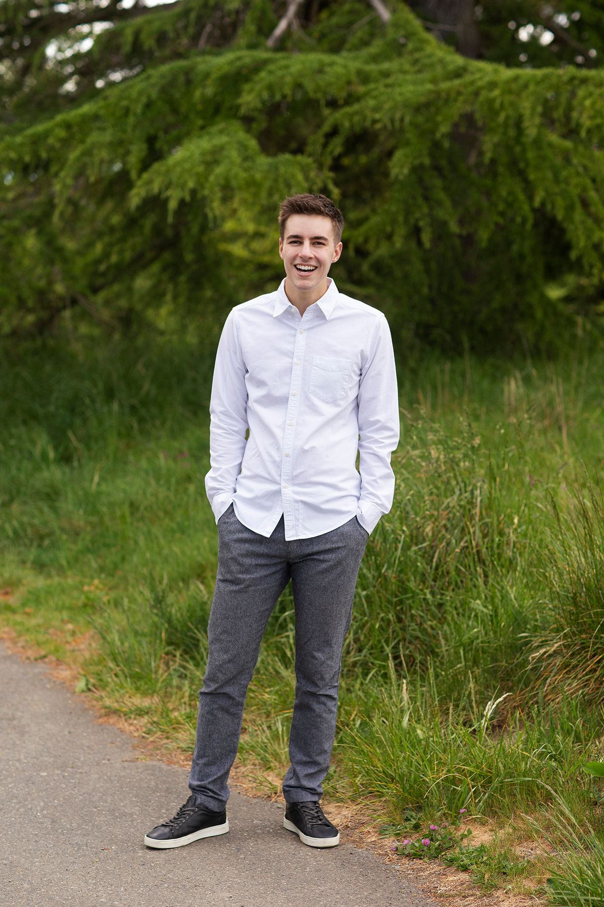 Young man in white shirt and gray pants smiling outdoors near green trees and grass.