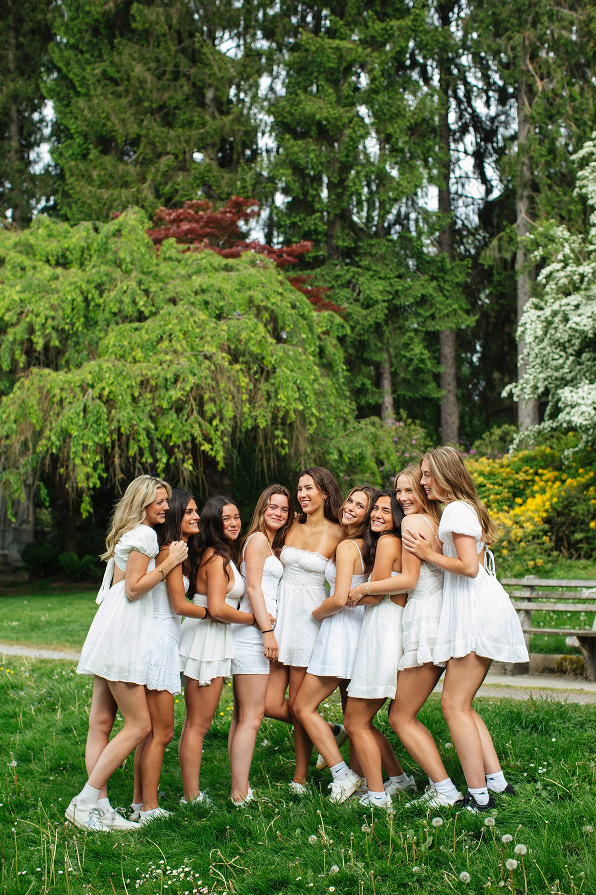 A group of ten young women in white dresses laugh and embrace together in a lush green park.