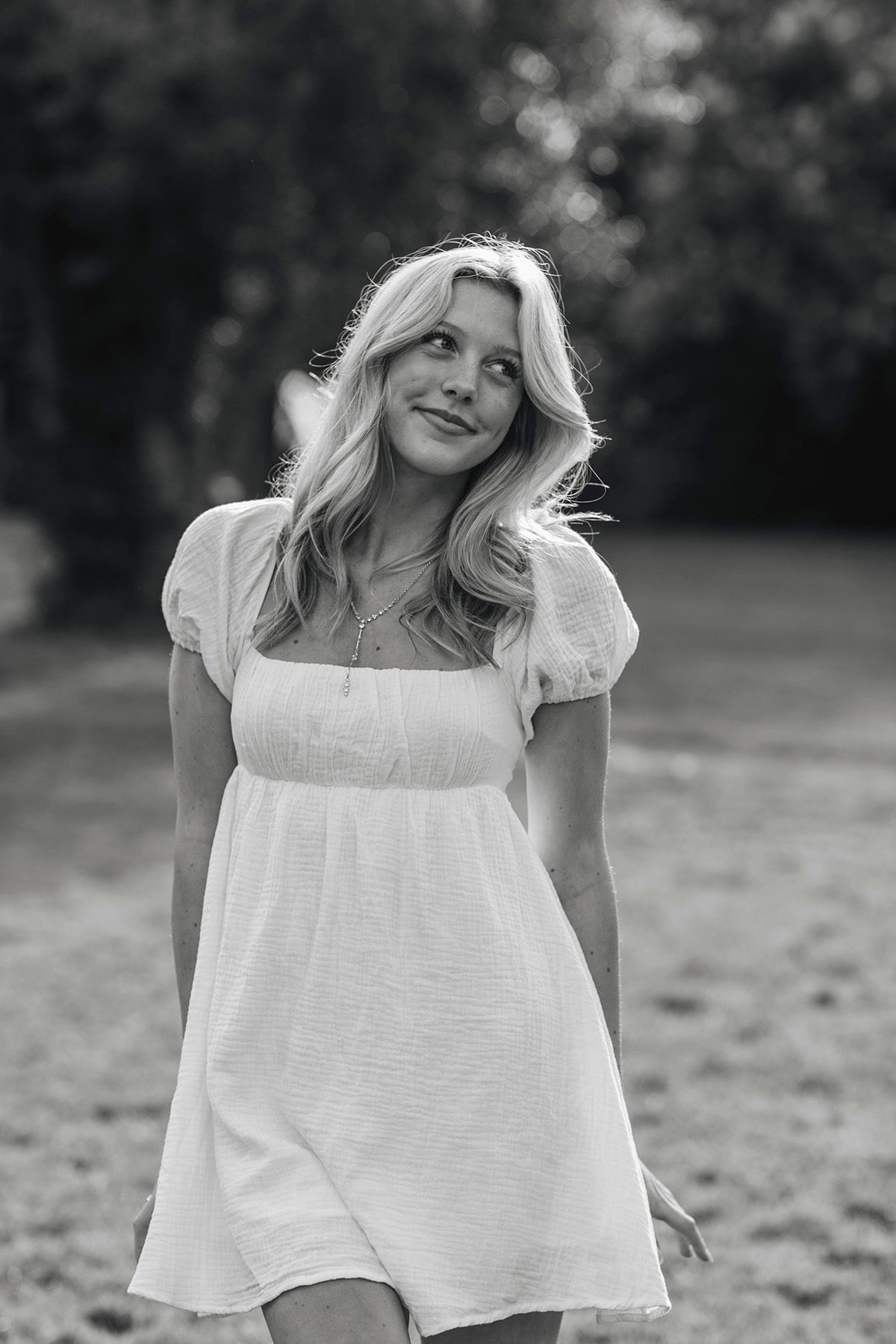 Black and white portrait of a young blonde woman in a white dress smiling outdoors in a garden setting.