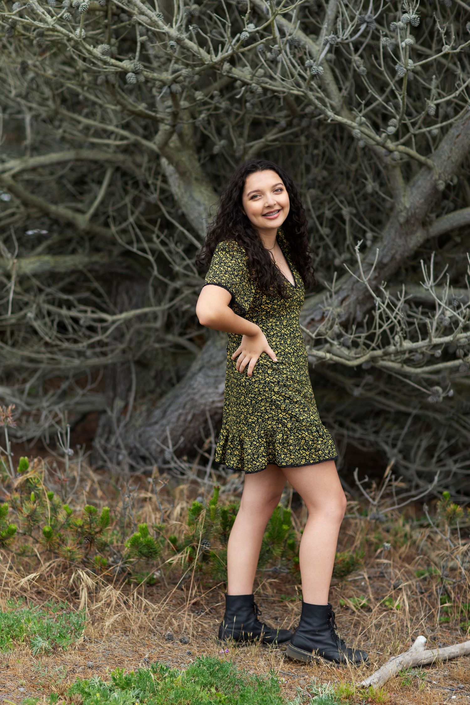 Young woman in green floral mini dress and black boots posing near bare trees.