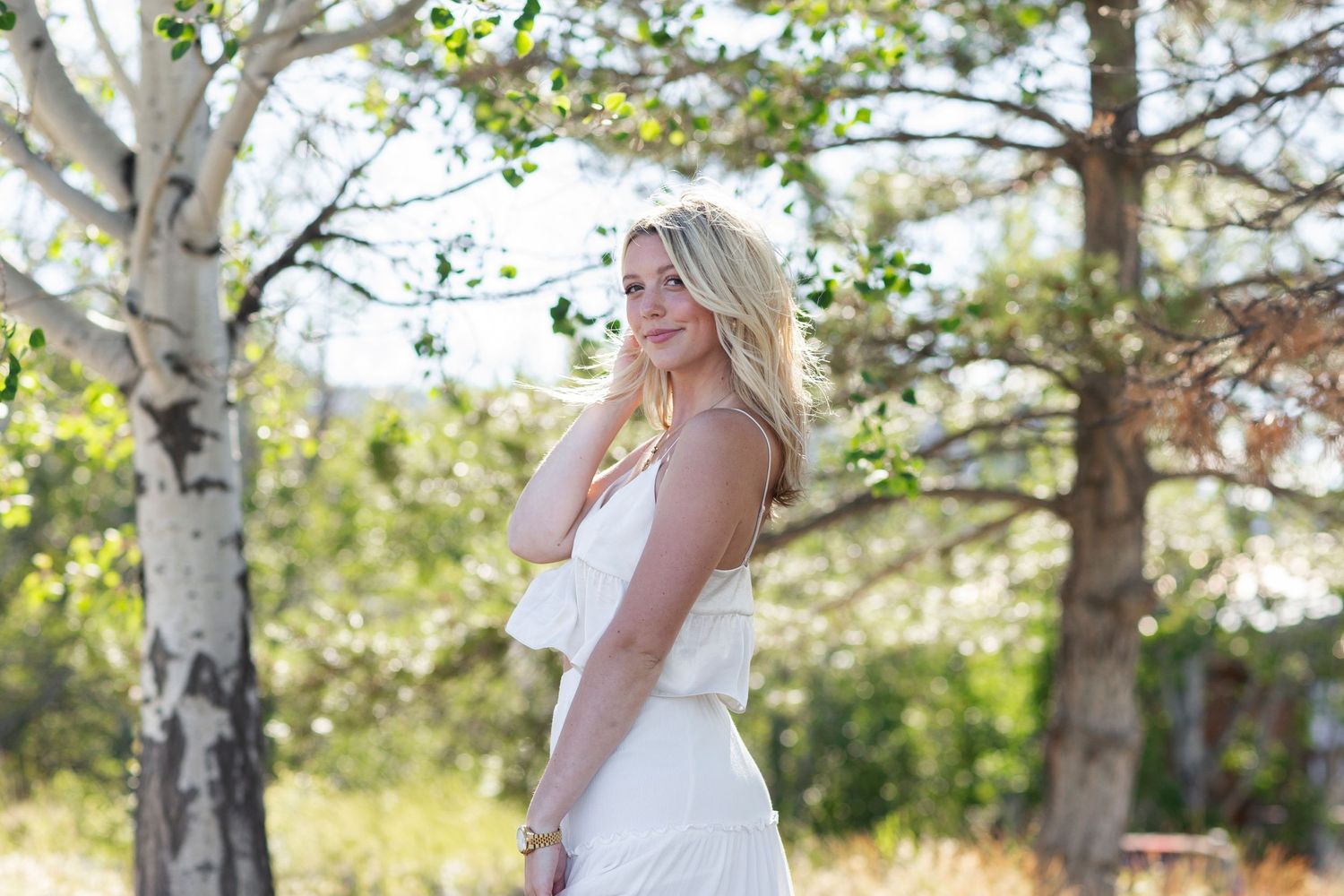 Young blonde woman in white dress posing gracefully among birch trees in a sunny outdoor setting.