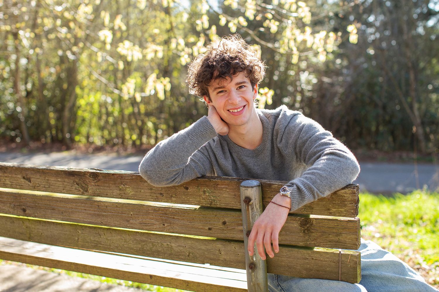 Smiling young man with curly hair leaning on a wooden park bench outdoors on a sunny day.
