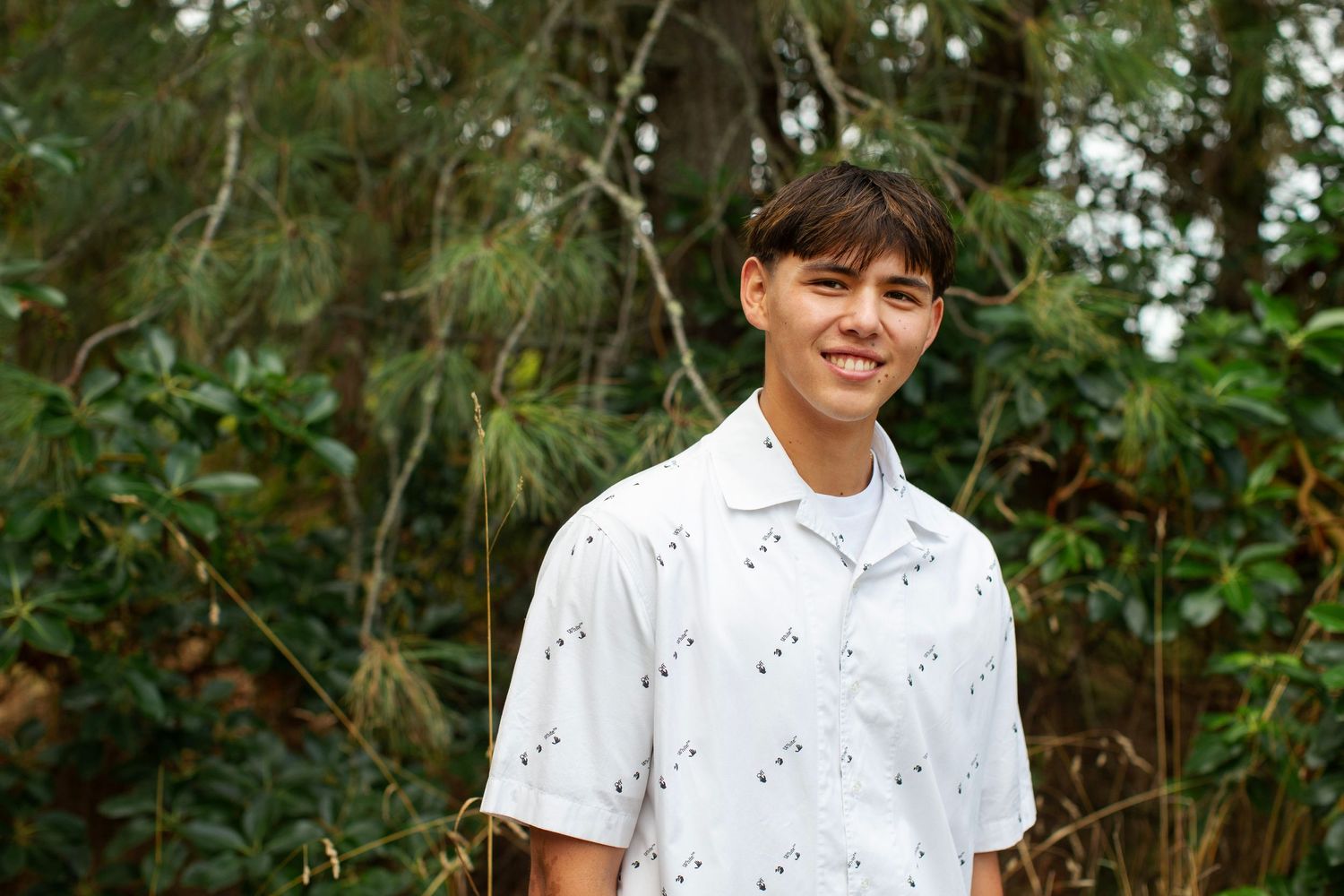 Smiling young man in white patterned shirt posing outdoors among lush green trees.