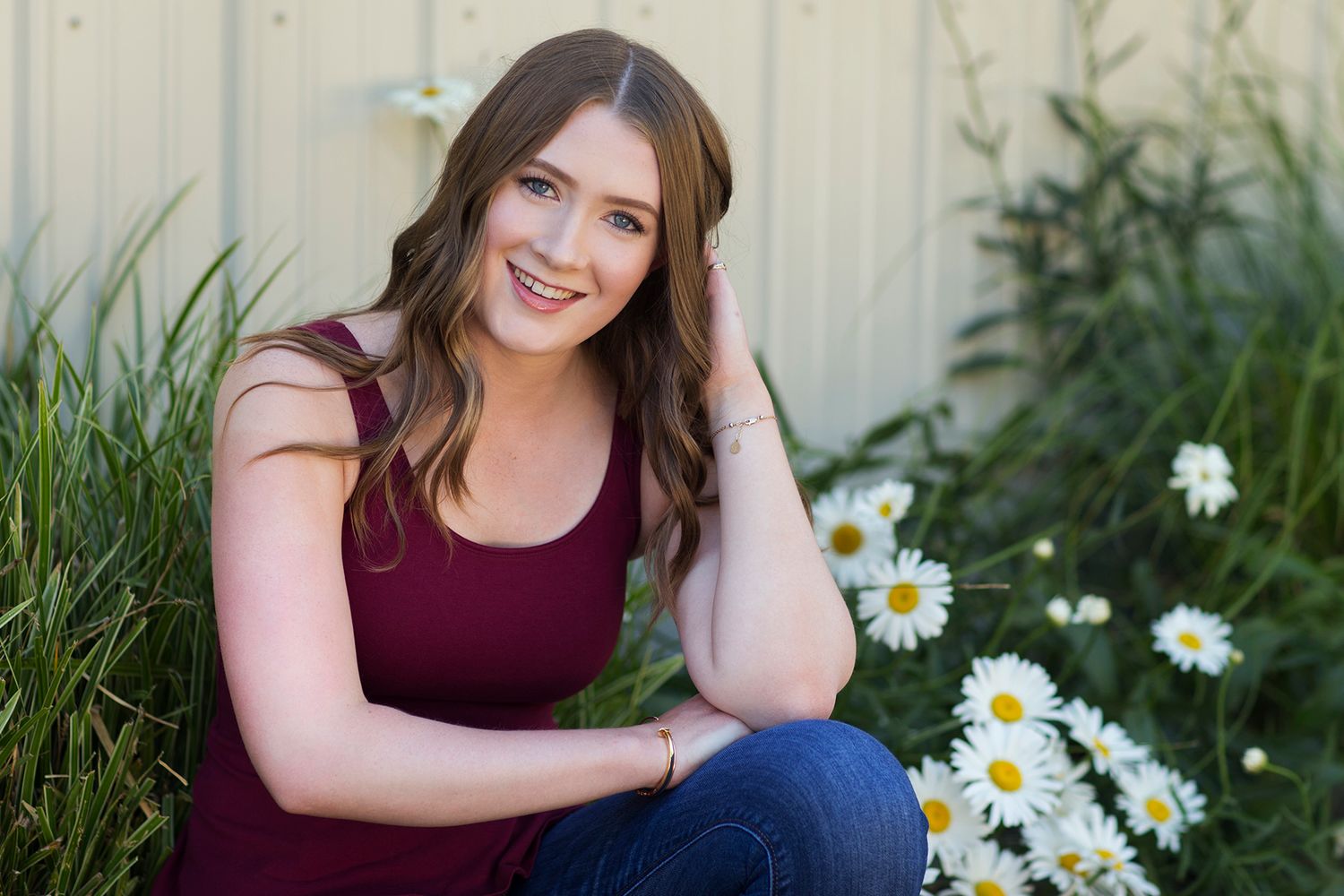 Young woman in burgundy top smiling outdoors beside white daisies, seated near a wooden fence.