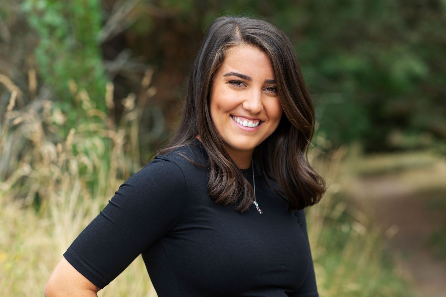Smiling brunette woman in black top posing outdoors on a nature trail with golden foliage.