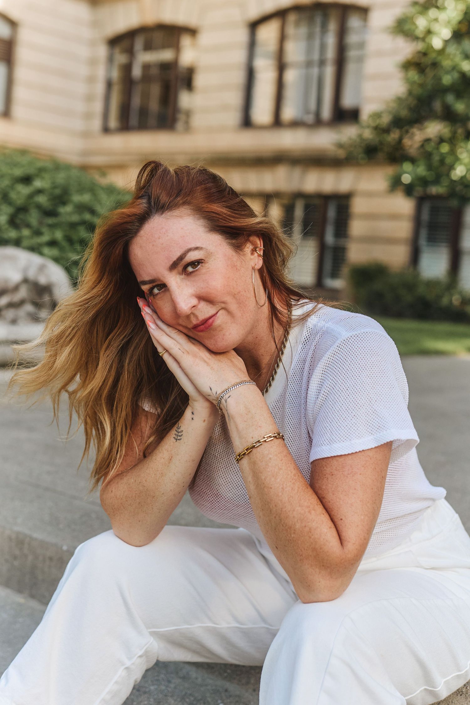 Smiling redhead woman in white outfit sitting outdoors near historic building in warm sunlight.