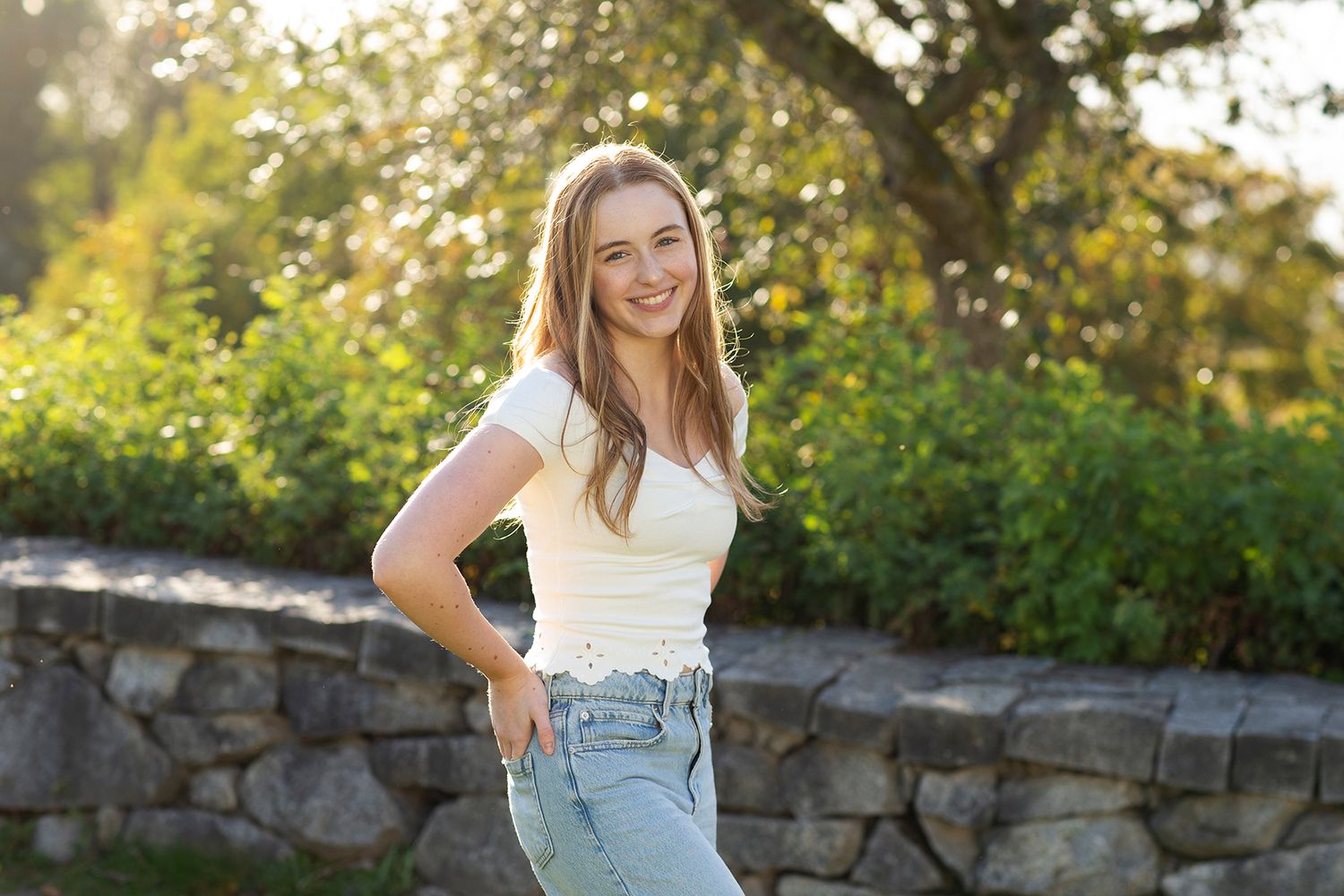 Young woman in white crop top and jeans smiling outdoors with lush green trees and stone wall background.