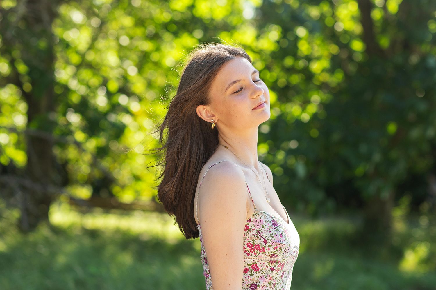 Young woman in floral dress enjoying sunlight outdoors in a lush green park on a sunny day.