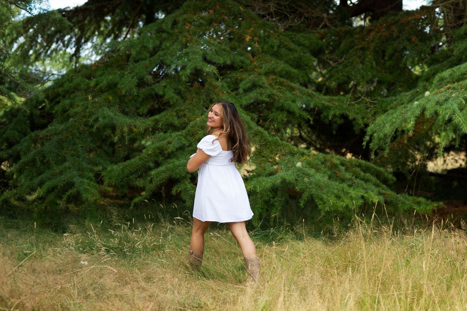 Young woman in a white dress laughing and twirling in a golden meadow in front of a large evergreen tree.