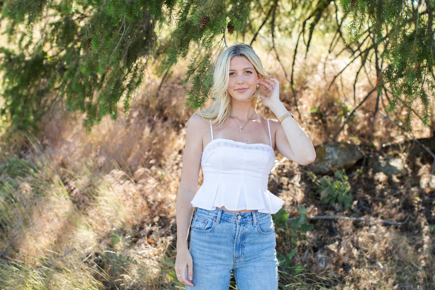 Smiling blonde woman in white cami top and jeans posing near evergreen trees in natural outdoor setting.