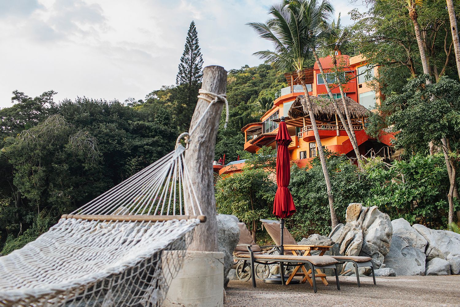 Hammock and lounge chairs on a rocky beach with a vibrant orange treehouse resort nestled in jungle trees.