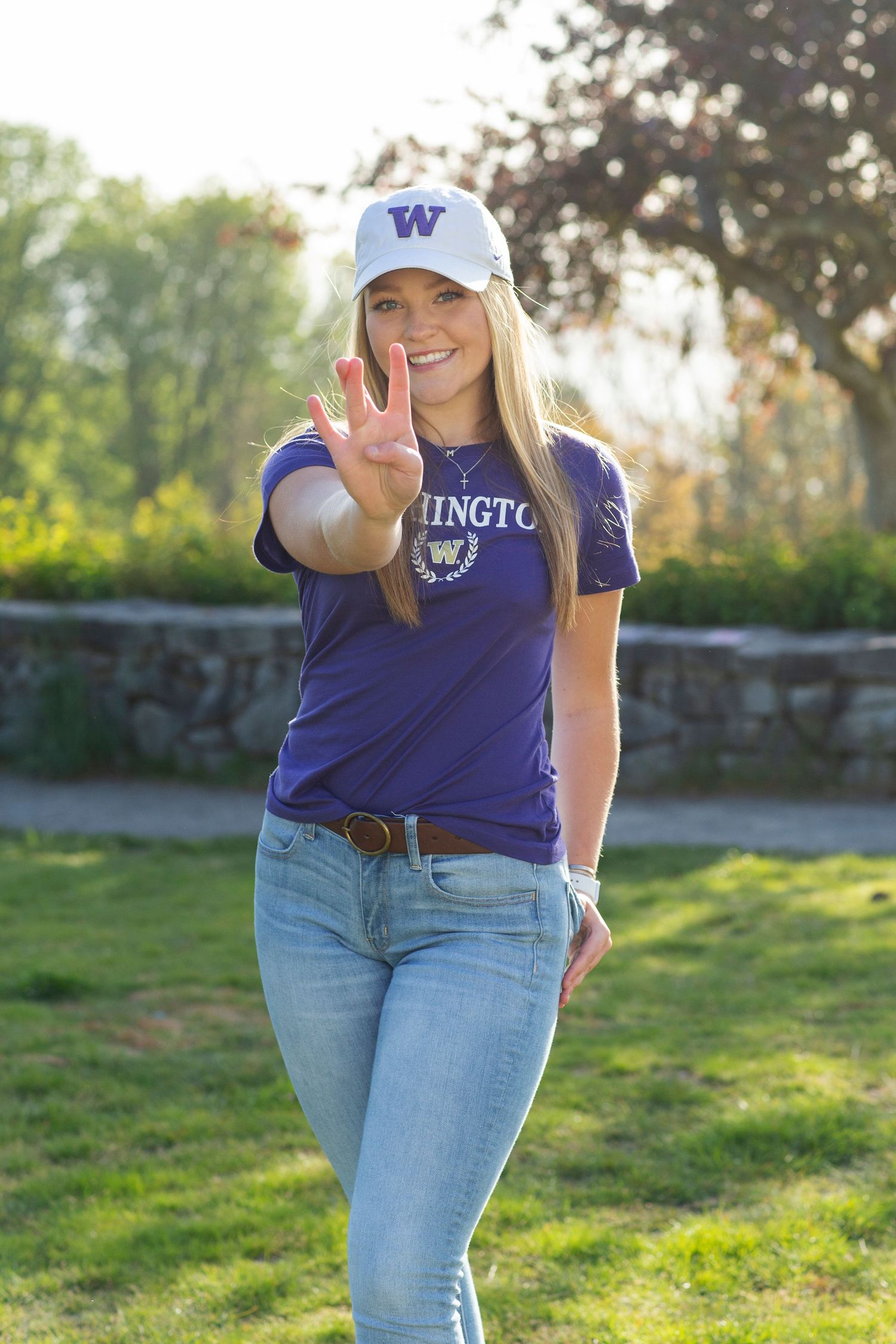 Young woman in University of Washington purple shirt and white W cap holding up three fingers outdoors.