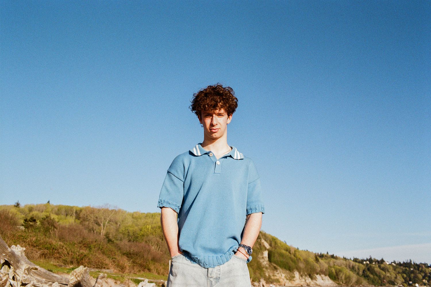 Young man in blue polo shirt standing outdoors against clear blue sky on rocky terrain.