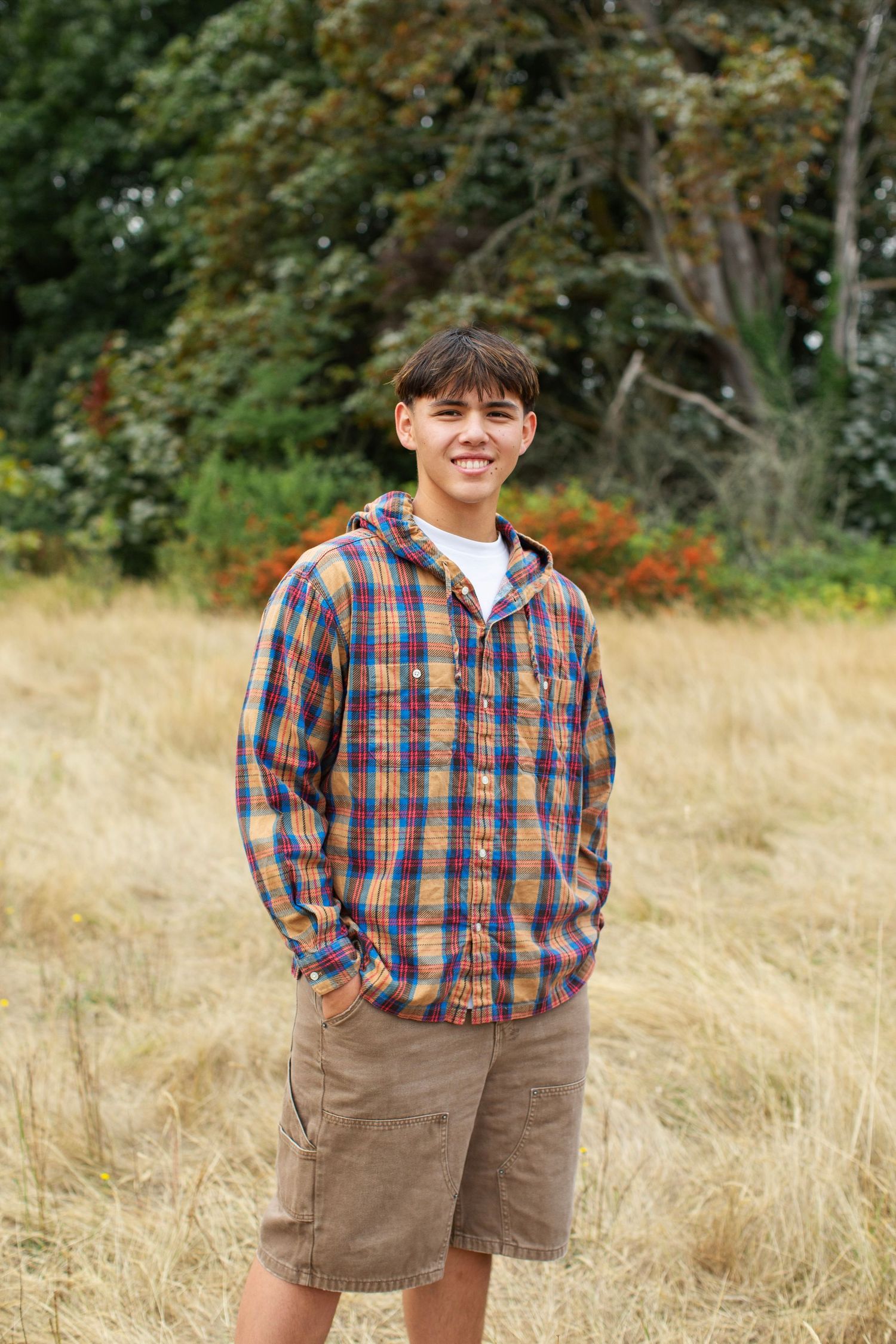 Teenage boy in colorful plaid shirt and khaki shorts standing in dry grass field.