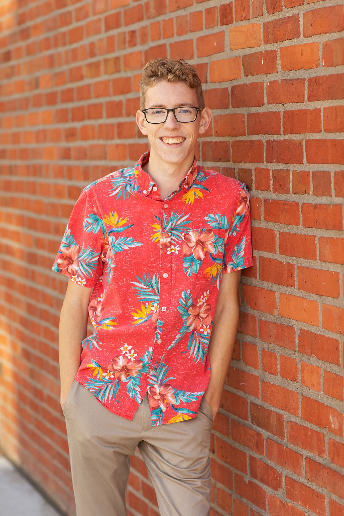 Smiling teen boy in red floral Hawaiian shirt and glasses leaning against a brick wall outdoors.