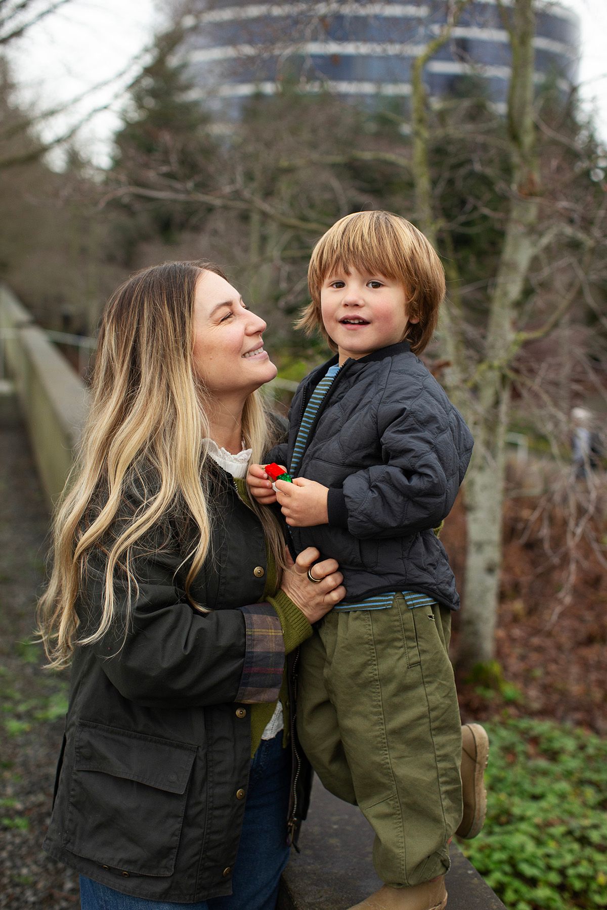 A woman with long blonde hair holds a smiling toddler boy outdoors in a park setting during winter.
