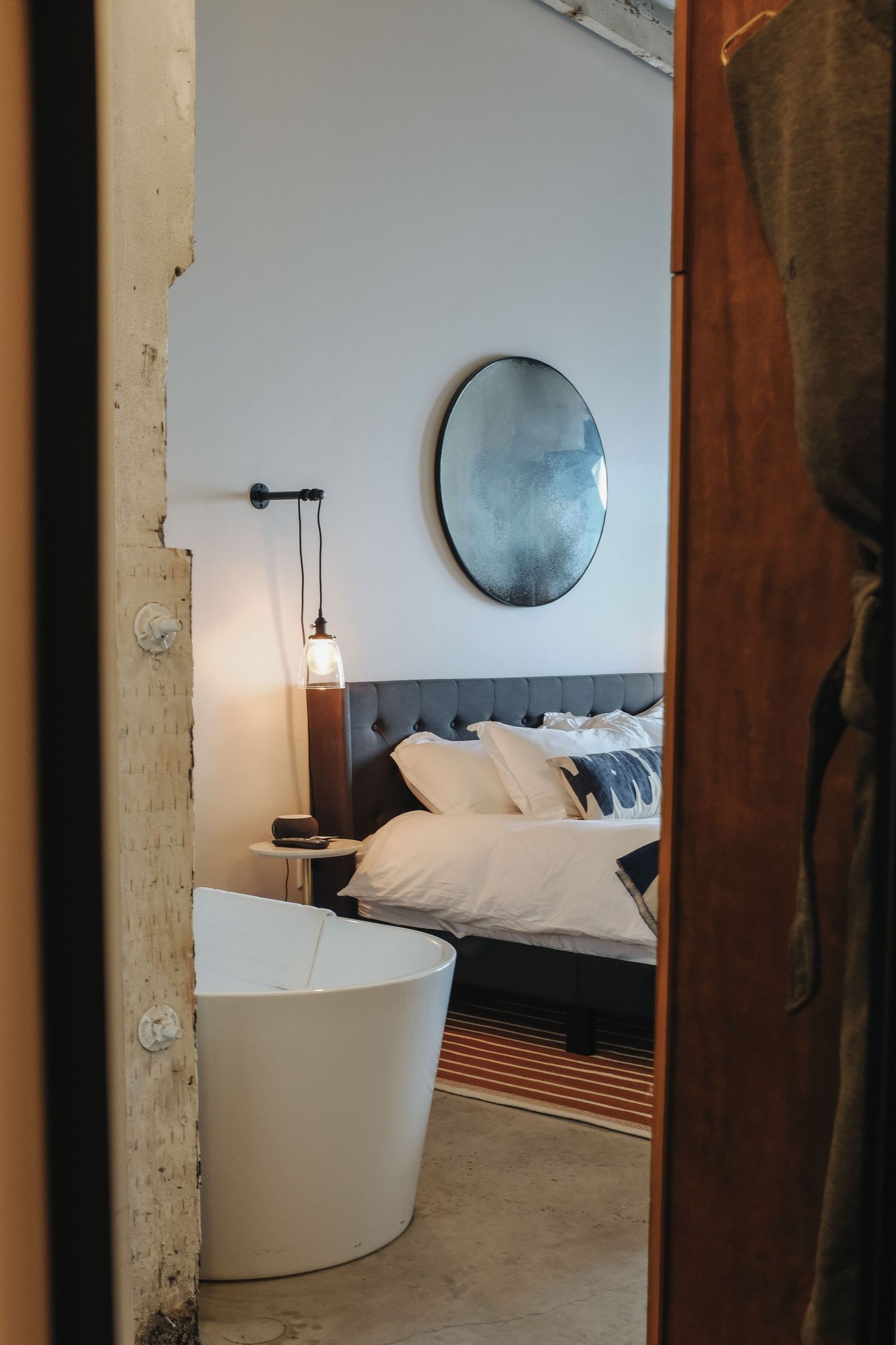 A cozy hotel room viewed through a doorway featuring a freestanding bathtub, pendant light, and plush bed.