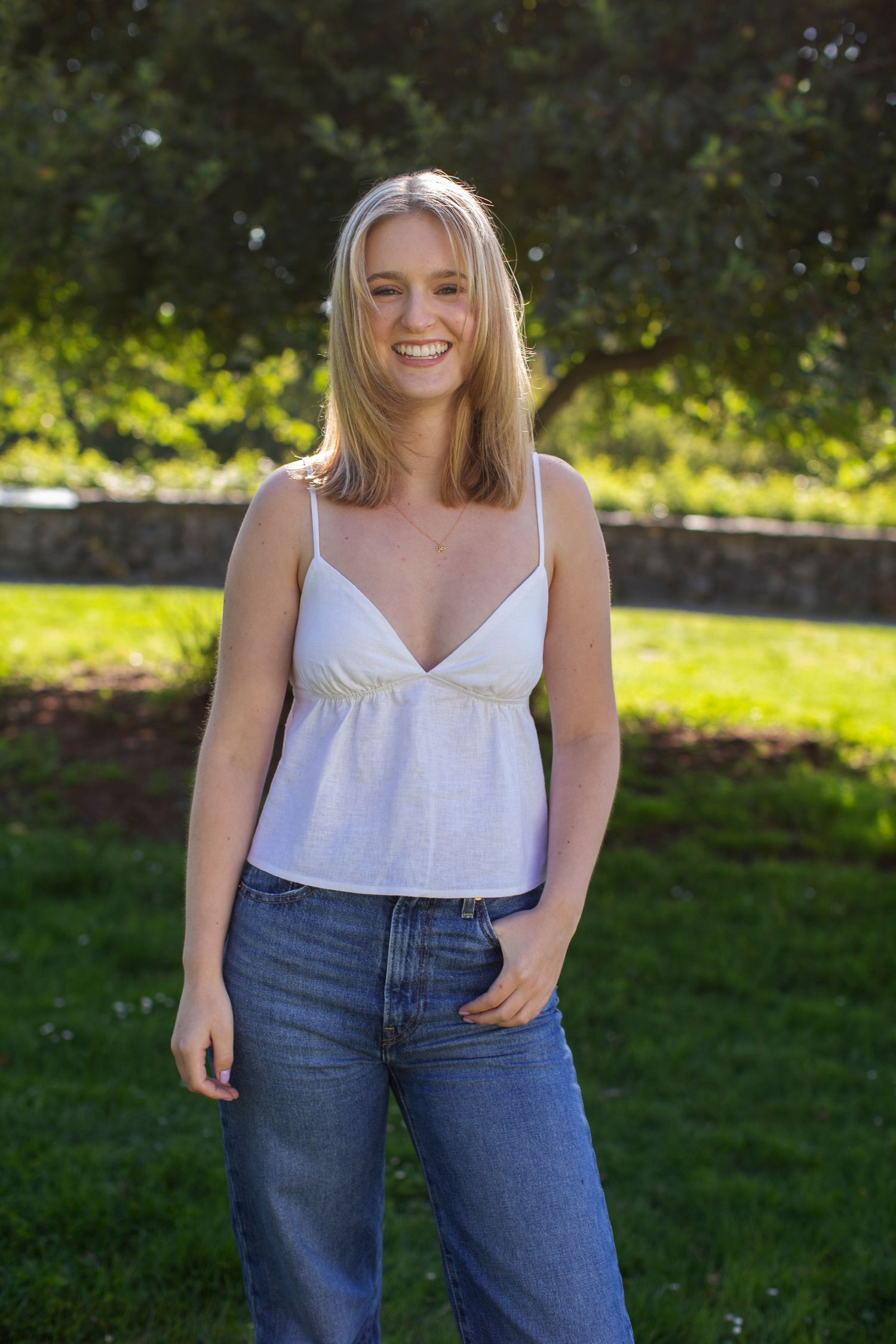 Happy blonde woman in white cami top and blue jeans smiling in sunny green park setting.