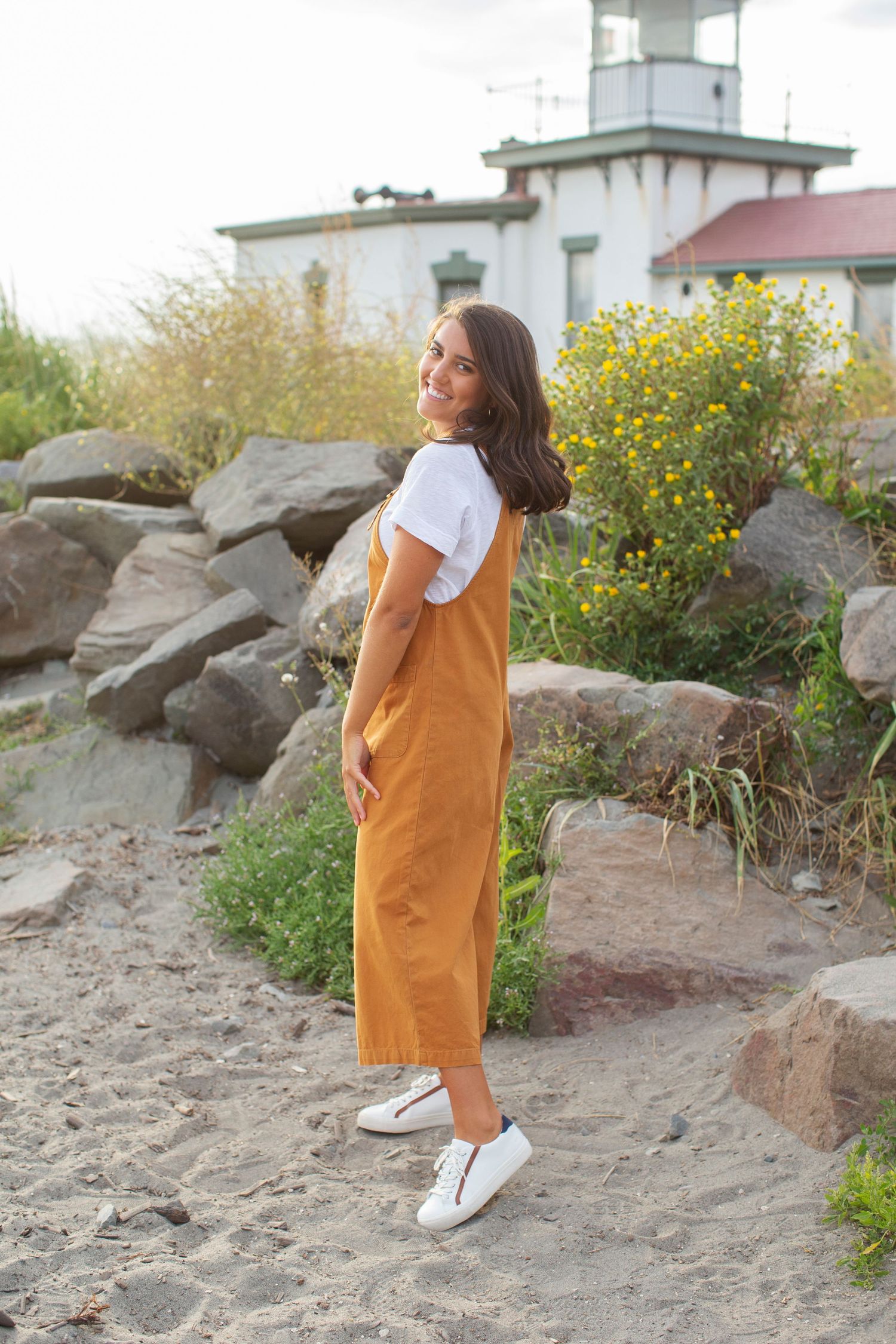 Senior Portrait Girl stands in front of lighthouse at Discovery Park with driftwood wearing orange overalls and sneakers at sunset