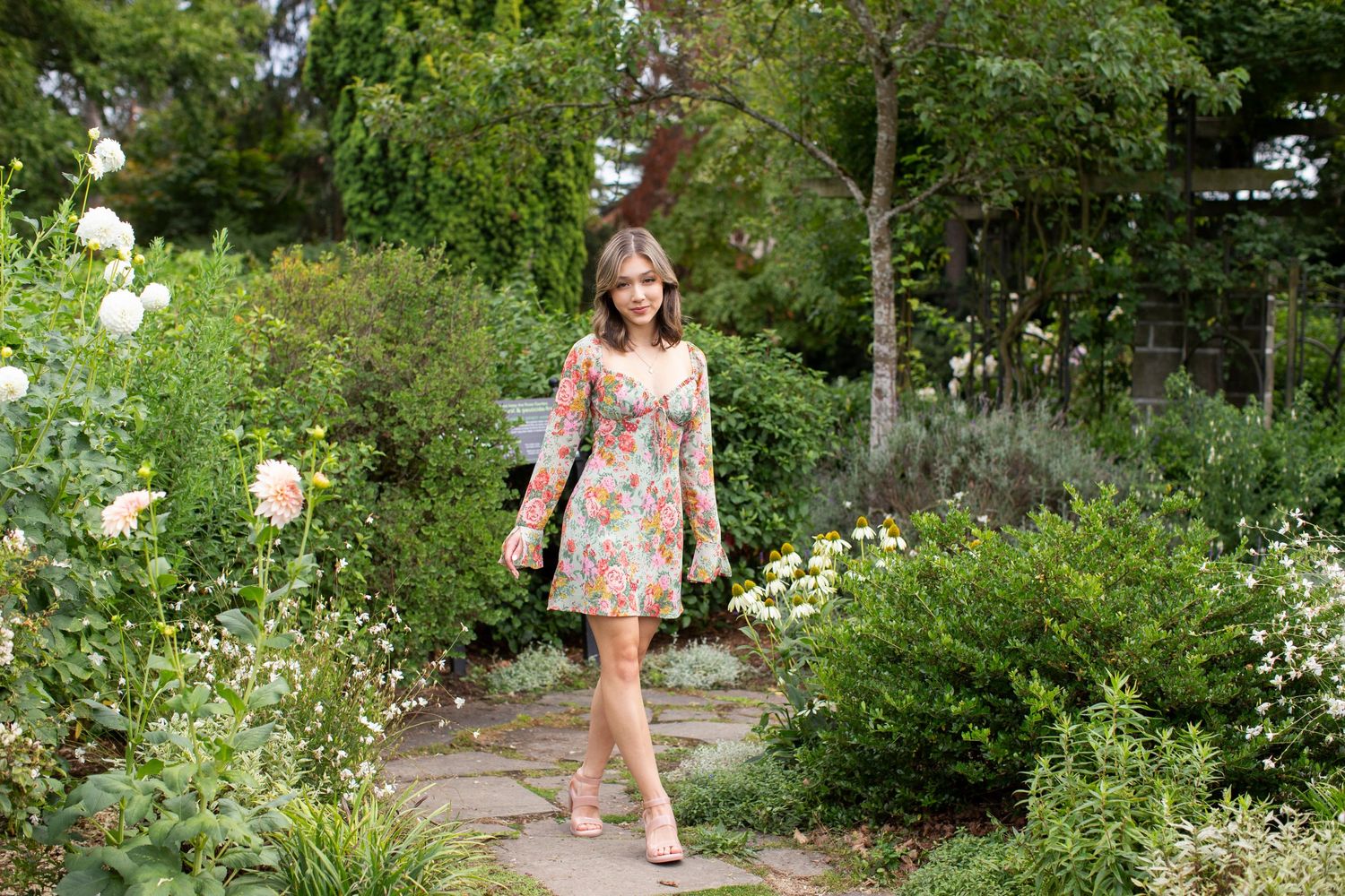 Young woman in a floral long-sleeve mini dress walking along a stone path in a lush garden.