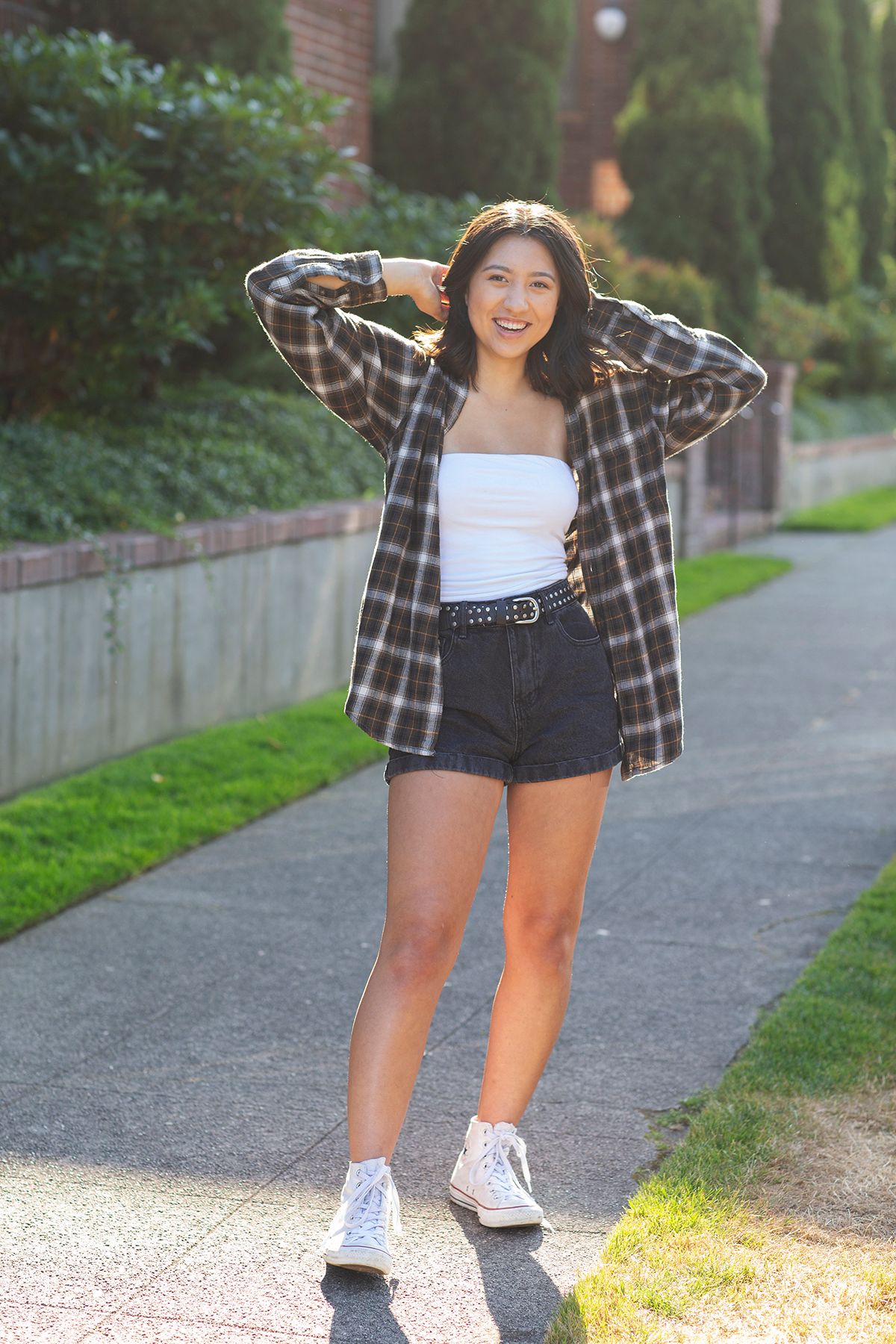 Young woman smiling in plaid shirt, white tube top, black denim shorts, and Converse sneakers, posing on a sunny sidewalk.