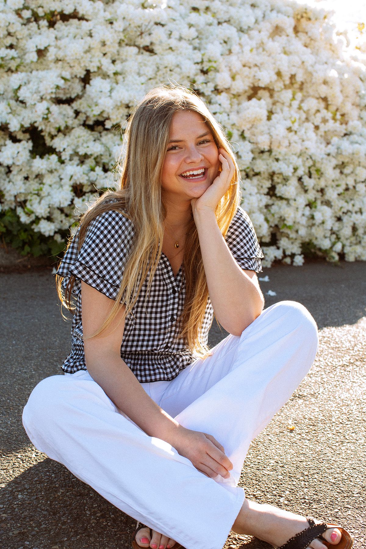 Smiling young woman in gingham top and white pants sitting cross-legged outdoors near white flowering bushes.