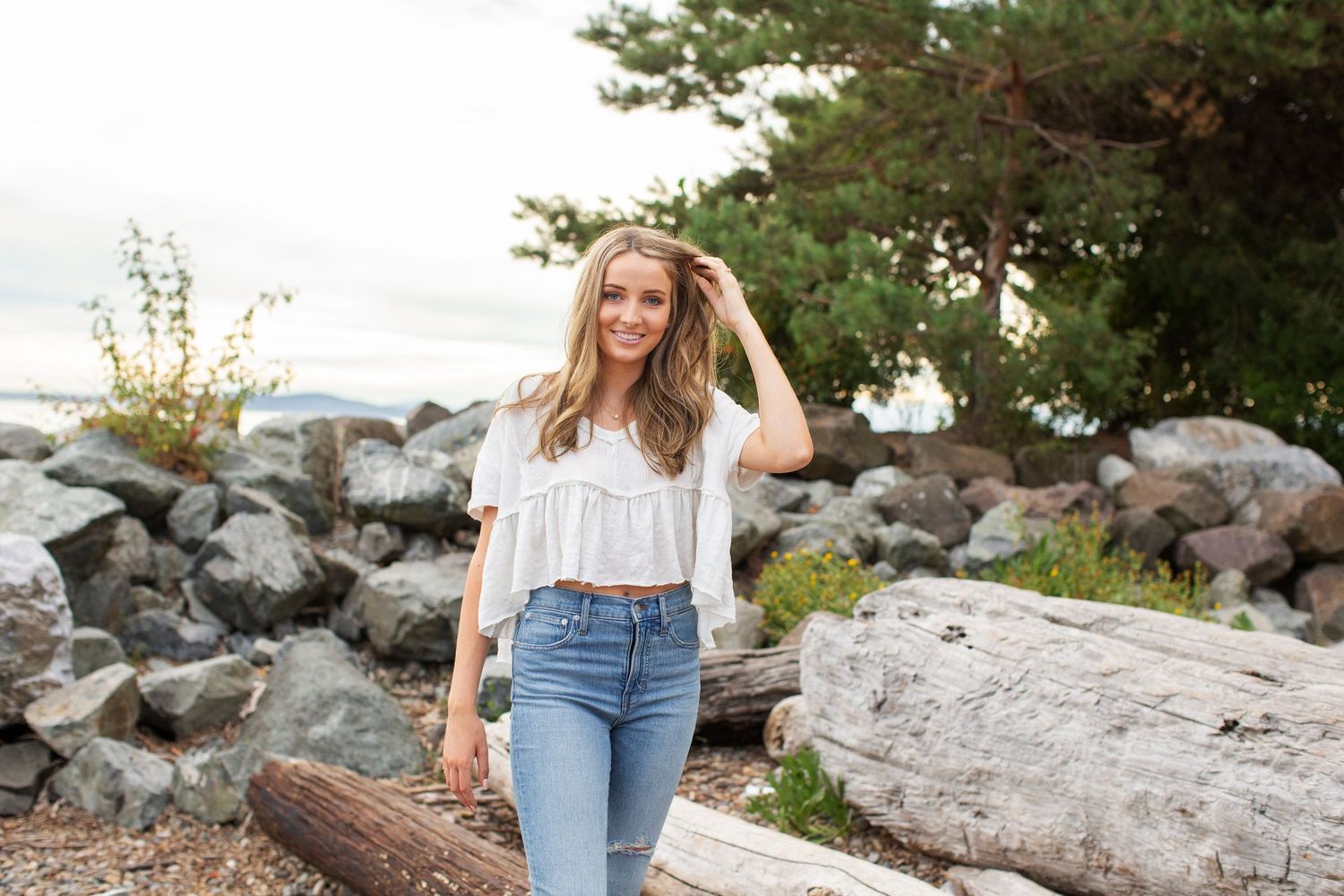 Smiling young woman in white ruffled top and jeans posing near rocks and driftwood.