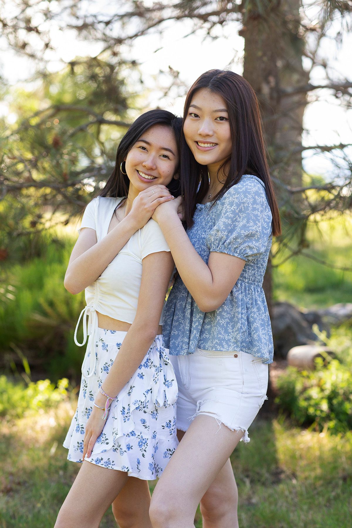 Two young Asian women smiling and posing together outdoors in summer outfits among green trees.