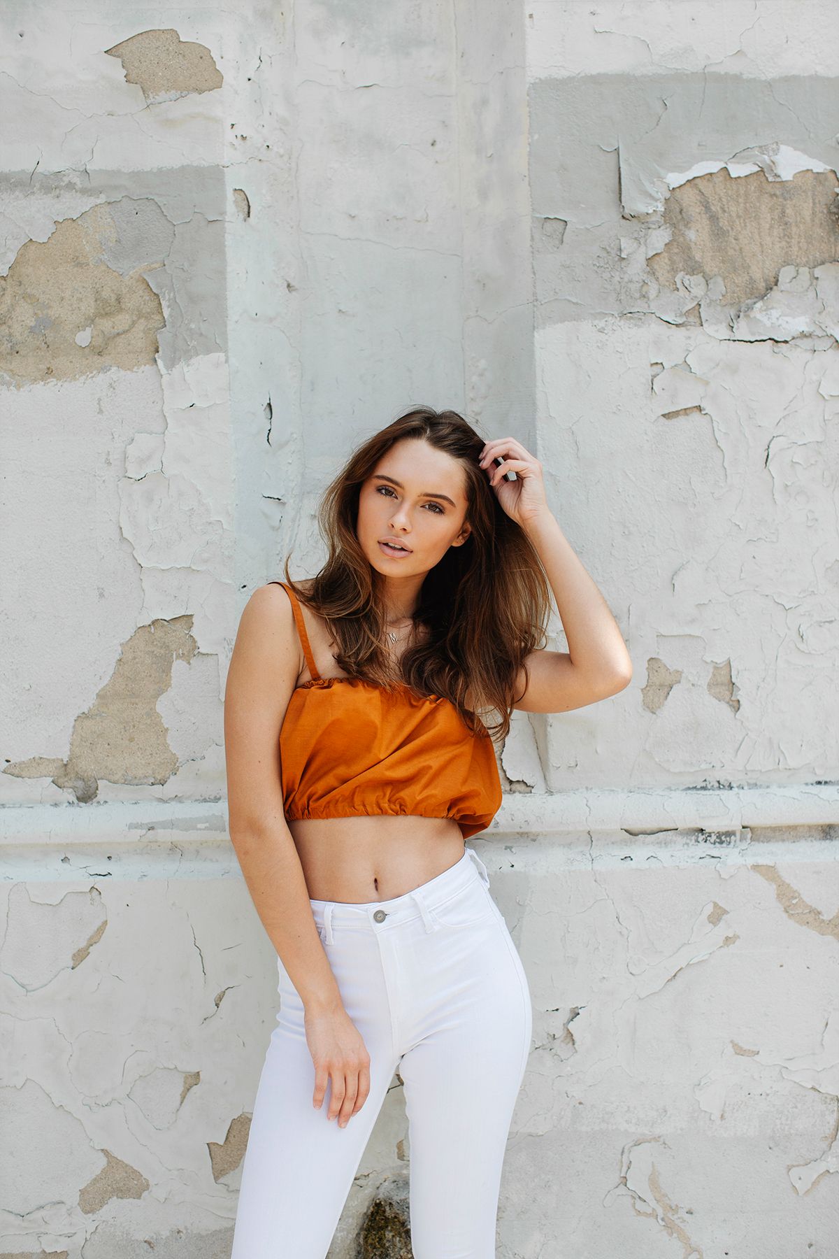 Young woman in orange crop top and white pants posing against distressed white wall with peeling paint.