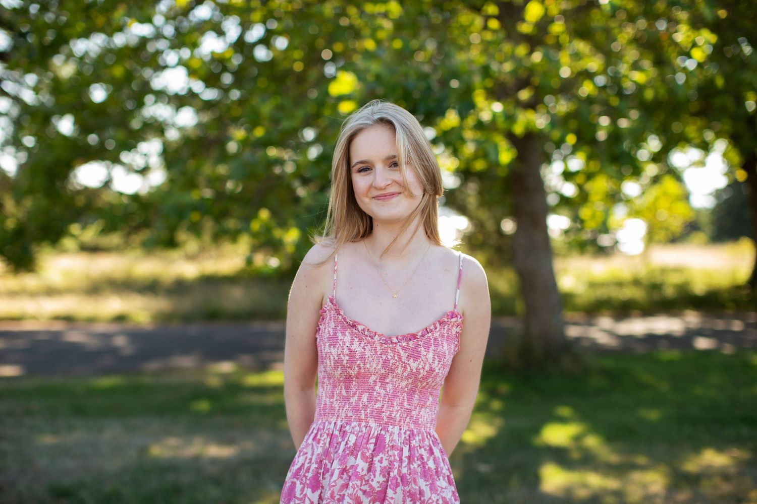 Young blonde woman in pink floral sundress smiling softly under green trees in summer park.