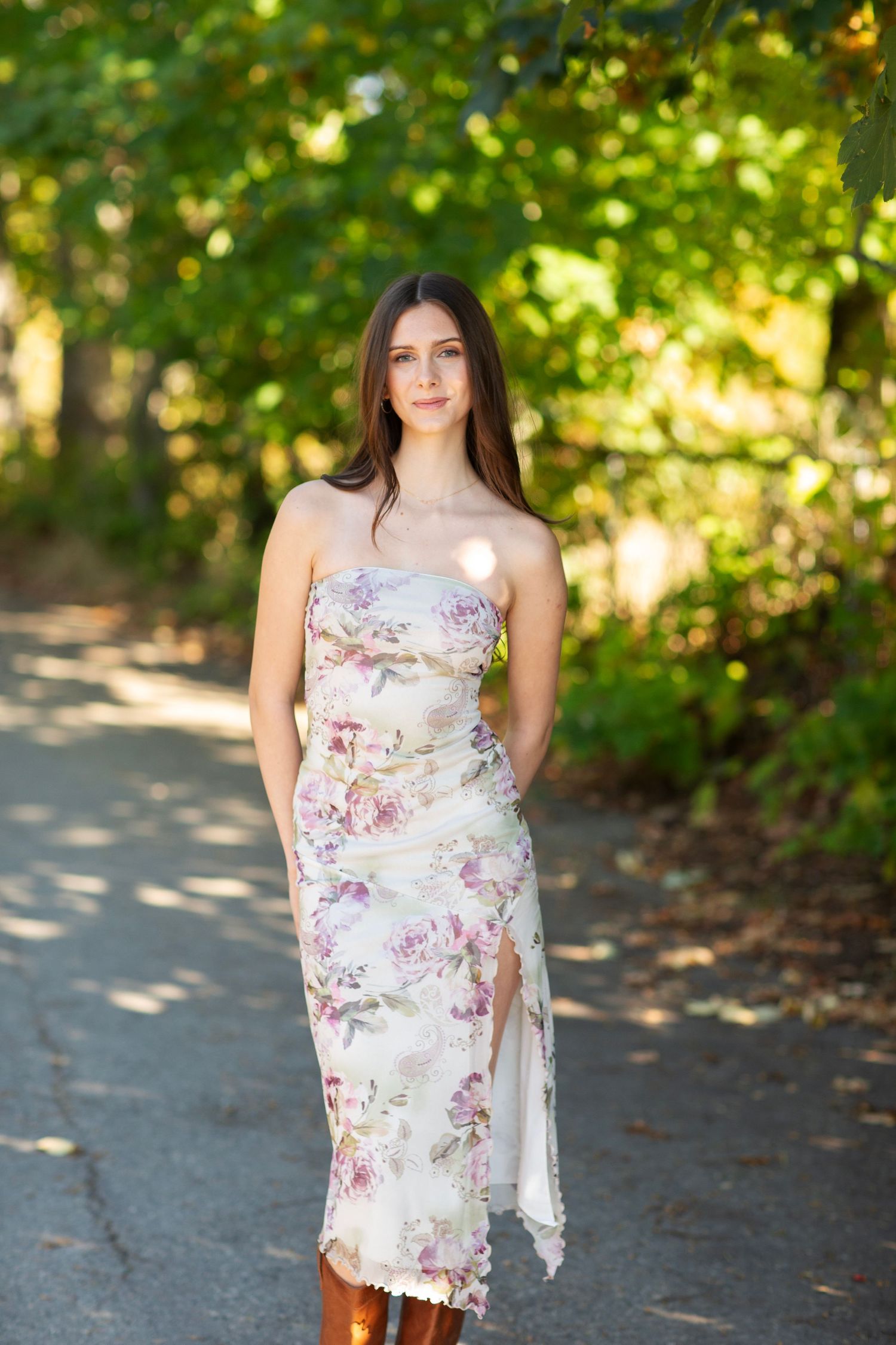 Young woman in strapless floral midi dress standing on tree-lined path in natural light.
